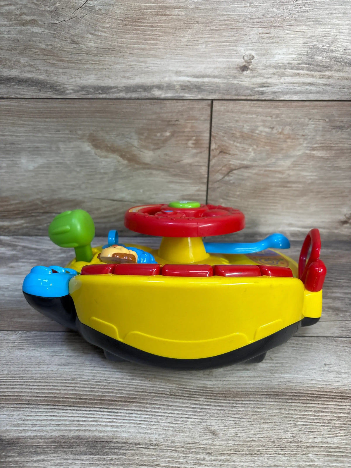 Side view of VTech Turn and Learn Driver toy showing red steering wheel and colorful buttons on yellow base on wood surface