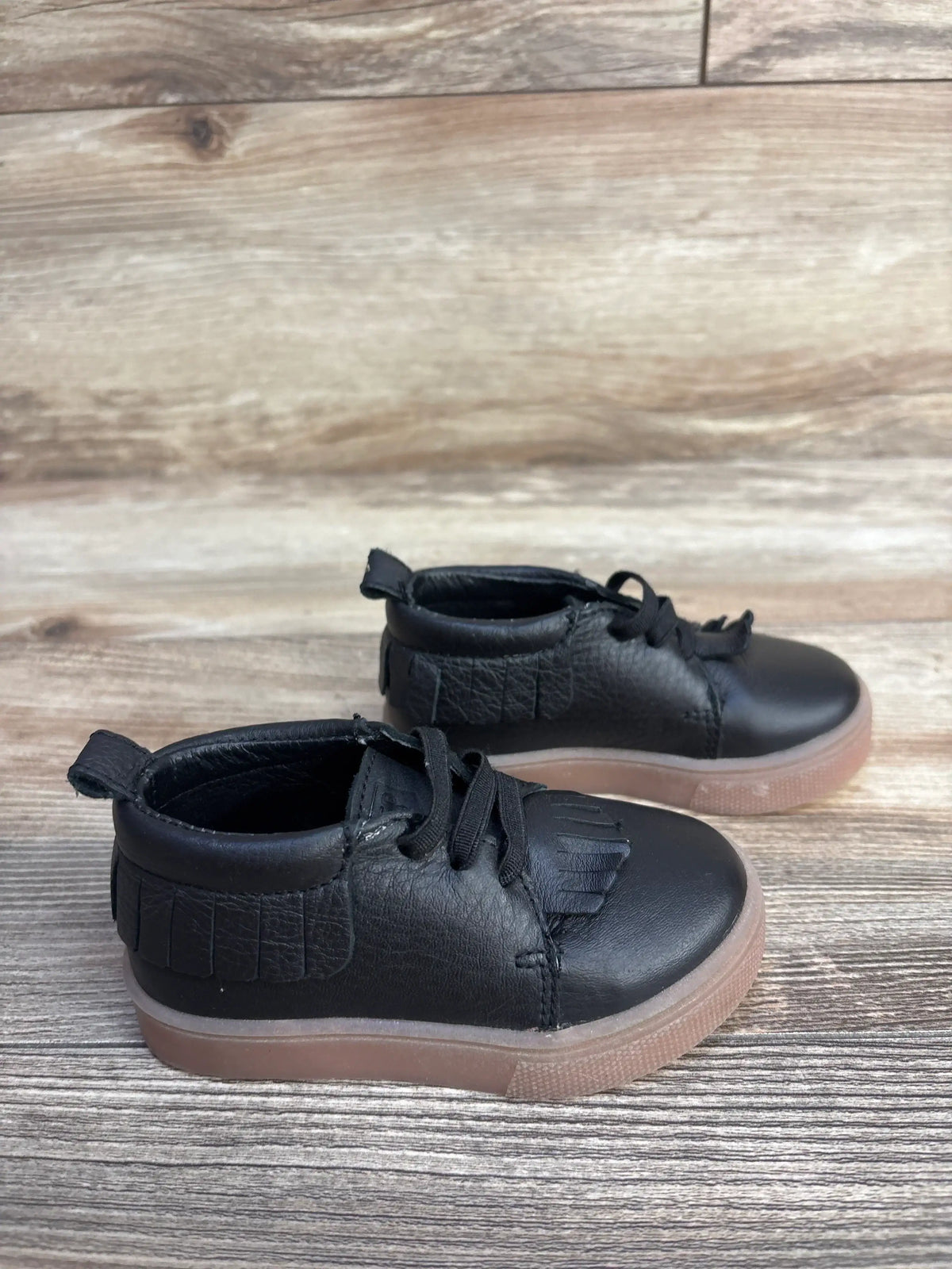 Side view of Freshly Picked black leather toddler slip-on mid sneakers showing fringe kiltie and gum sole on wood background