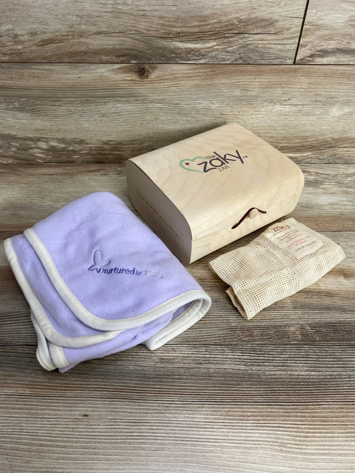 Angled view of Zaky Zak swaddle set: lavender swaddle with cream trim and nurtured by design embroidery, wooden keepsake box with logo, and mesh wash bag on wood