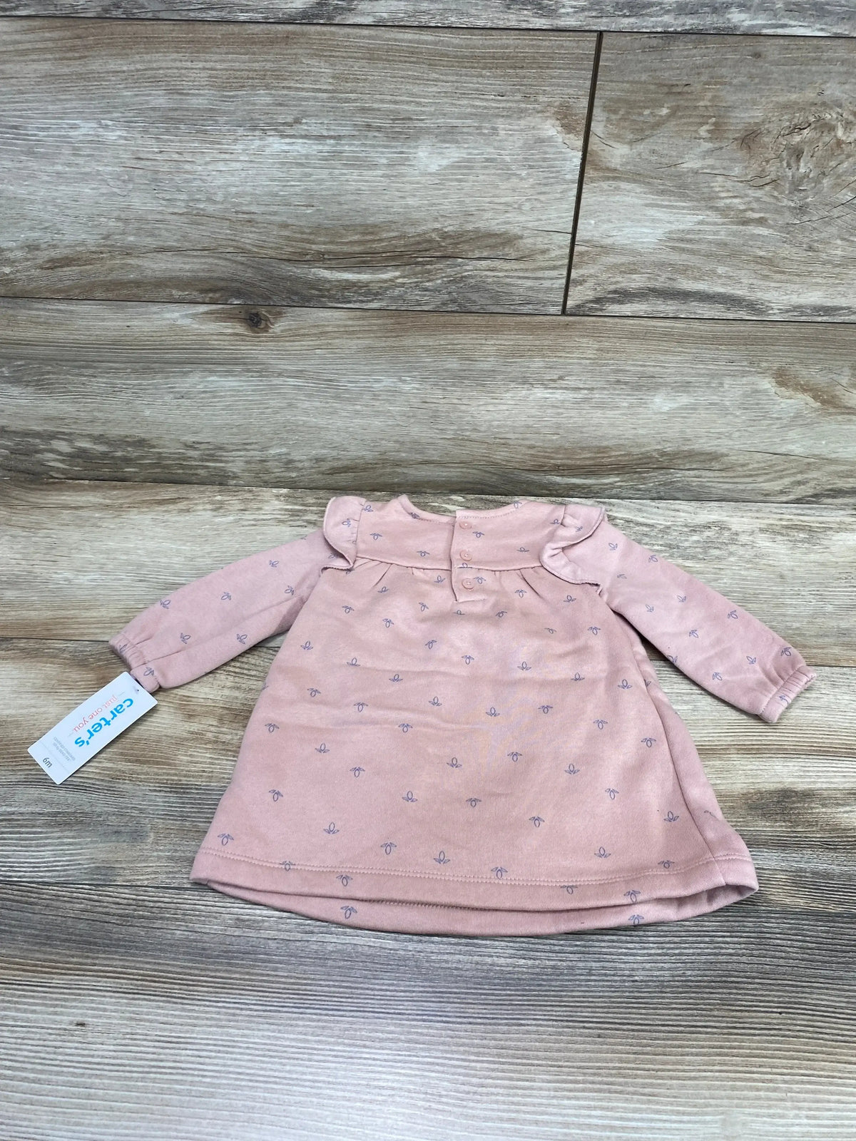 Flat lay of pink baby sweatshirt dress showing ruffle collar, long sleeves, and small purple bow print on wood background