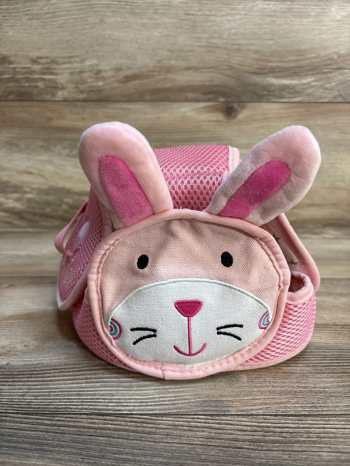 Front of Jjovce baby safety hat, pink bunny, showing plush ears, embroidered face, mesh padding and chin strap on wood surface