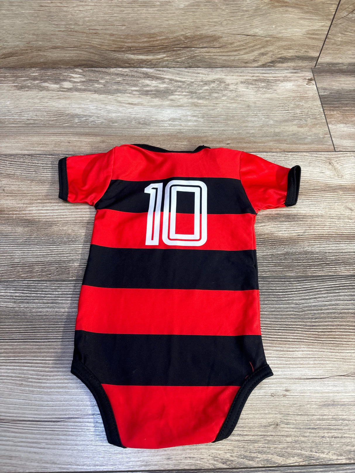 Flat lay of Flamengo baby bodysuit, red and black stripes with white 10 on back, on light wood floor, size 0–3 months
