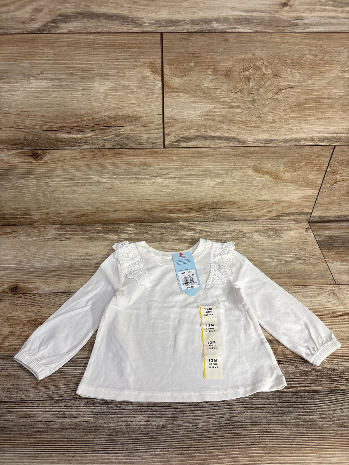 White infant long sleeve shirt with lace ruffle shoulders, size 12M, on wood background