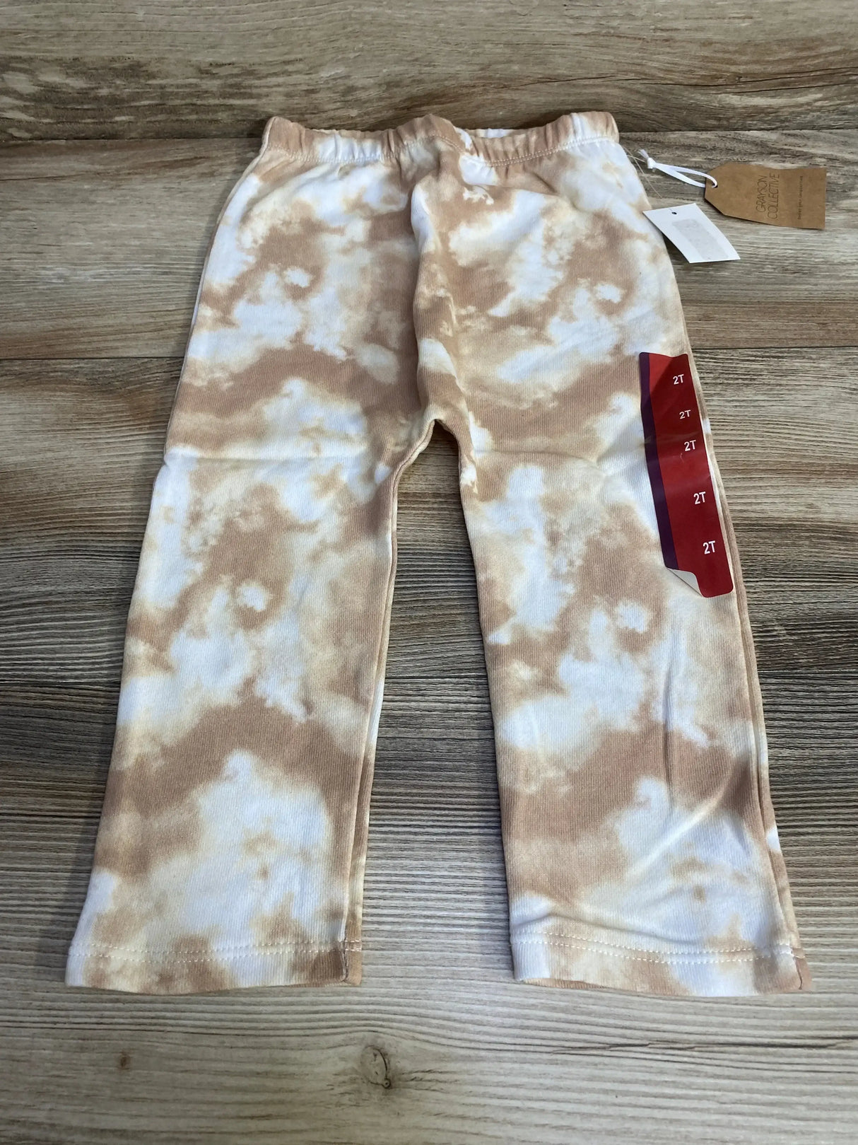 Top-down view of Grayson Collective tie-dye toddler pants, tan and white, elastic waistband, size 2T sticker on wood