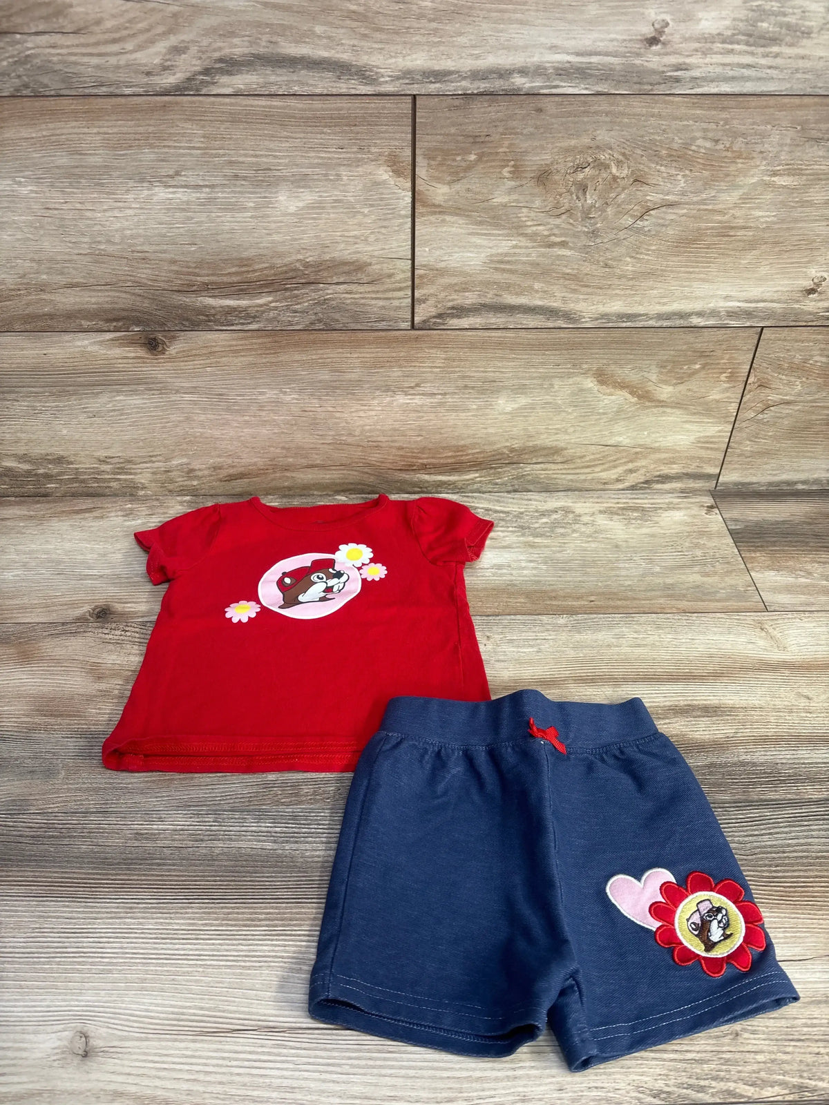 Flat lay of Buc-ees toddler 2-piece red t-shirt and navy shorts showing front beaver graphic on shirt and red flower patch on shorts on wood floor background