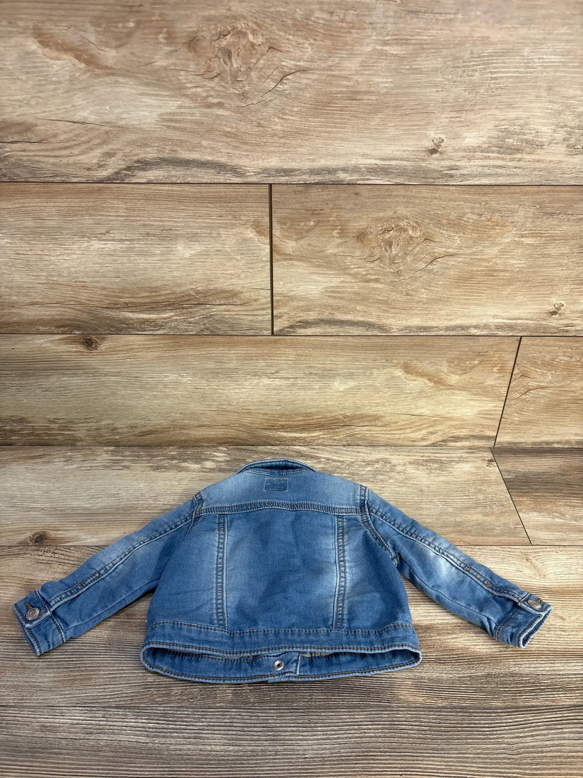 Back view of OshKosh blue denim jacket, size 12 months, showcasing panel seams and long sleeves on a wood floor