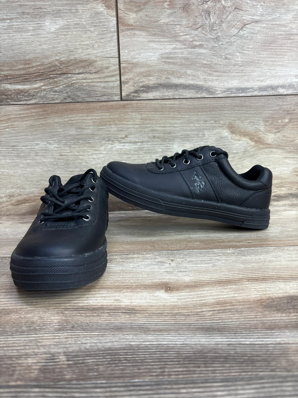 Black kids athletic shoes with laces, textured sides, and rubber soles on wooden floor