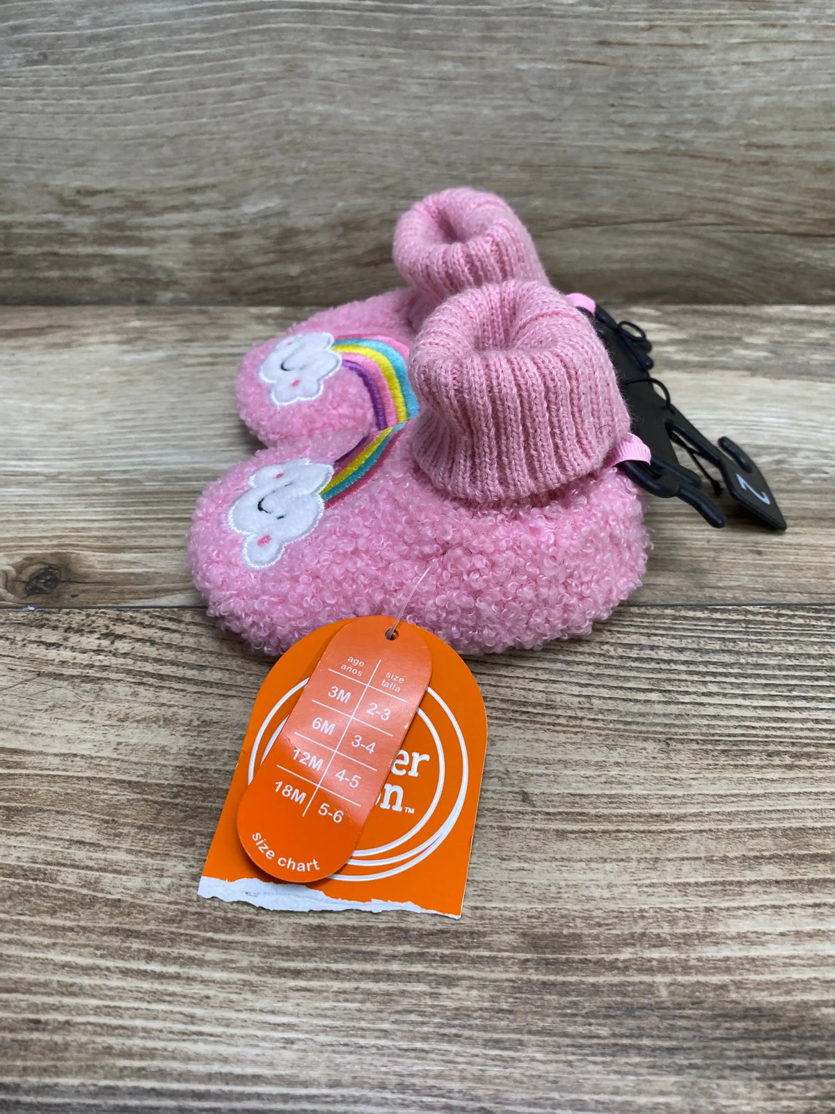 Angled view of pink baby bootie slippers with embroidered cloud and rainbow, knit cuff and fuzzy sole on wood background