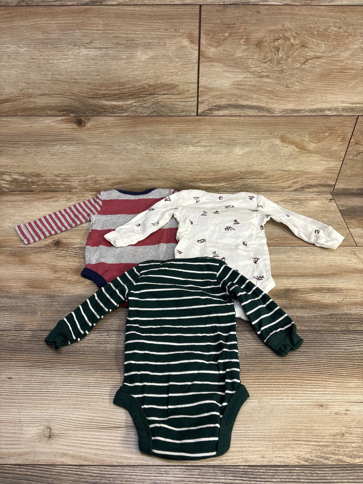 Three long-sleeve baby bodysuits on wooden floor, striped green, striped red-gray, and white with small prints