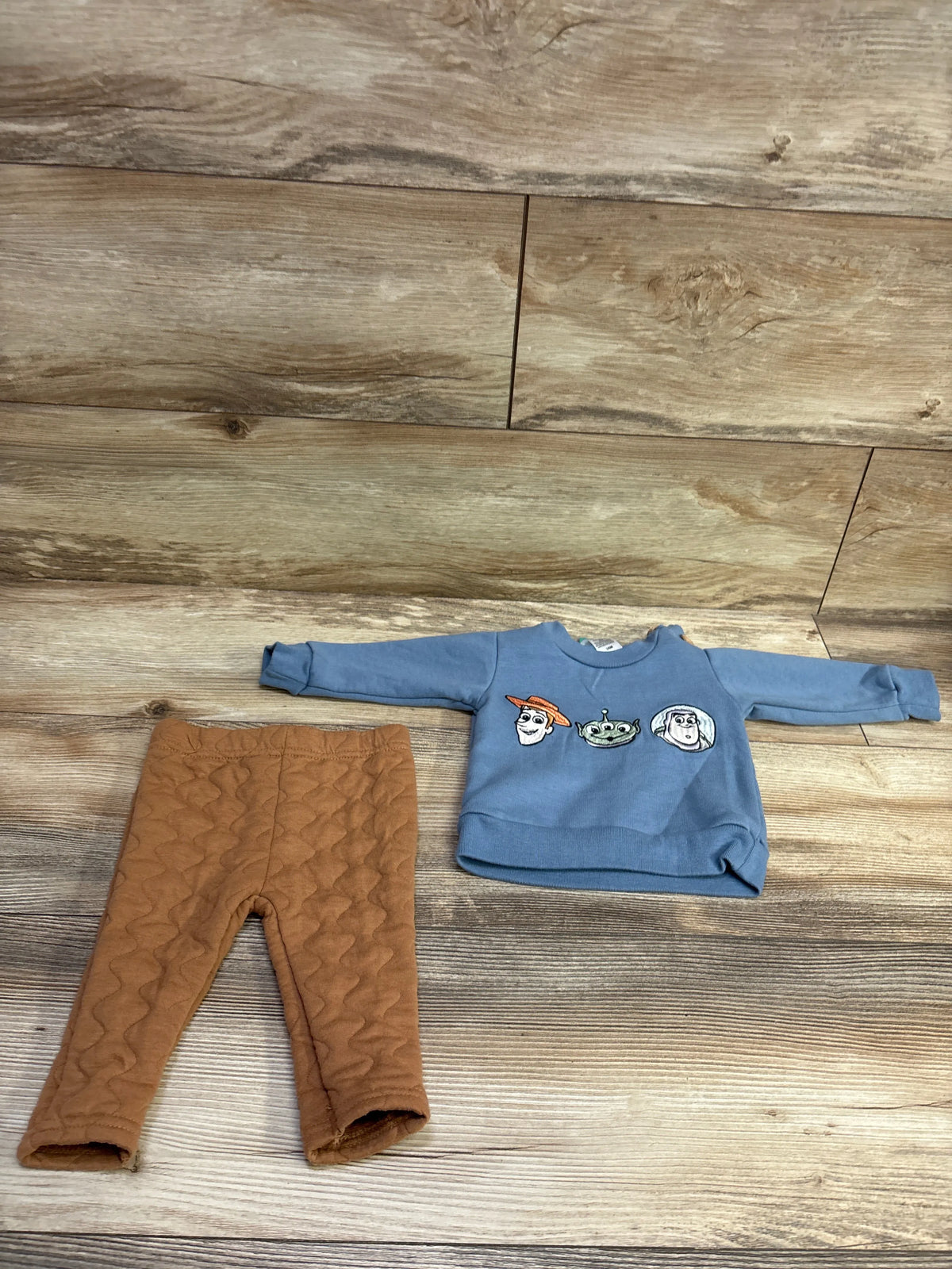 Flat lay of a Disney Baby Toy Story sweatshirt and pants set with embroidered character faces, blue sweatshirt, and brown quilted pants