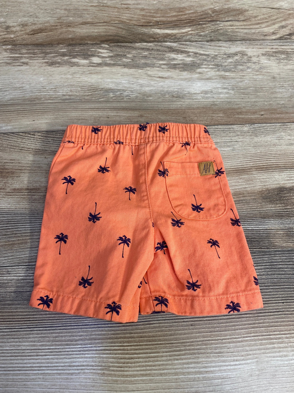 Back view of orange baby shorts size 6-9 months with navy palm print, elastic waistband and back patch pocket on wood