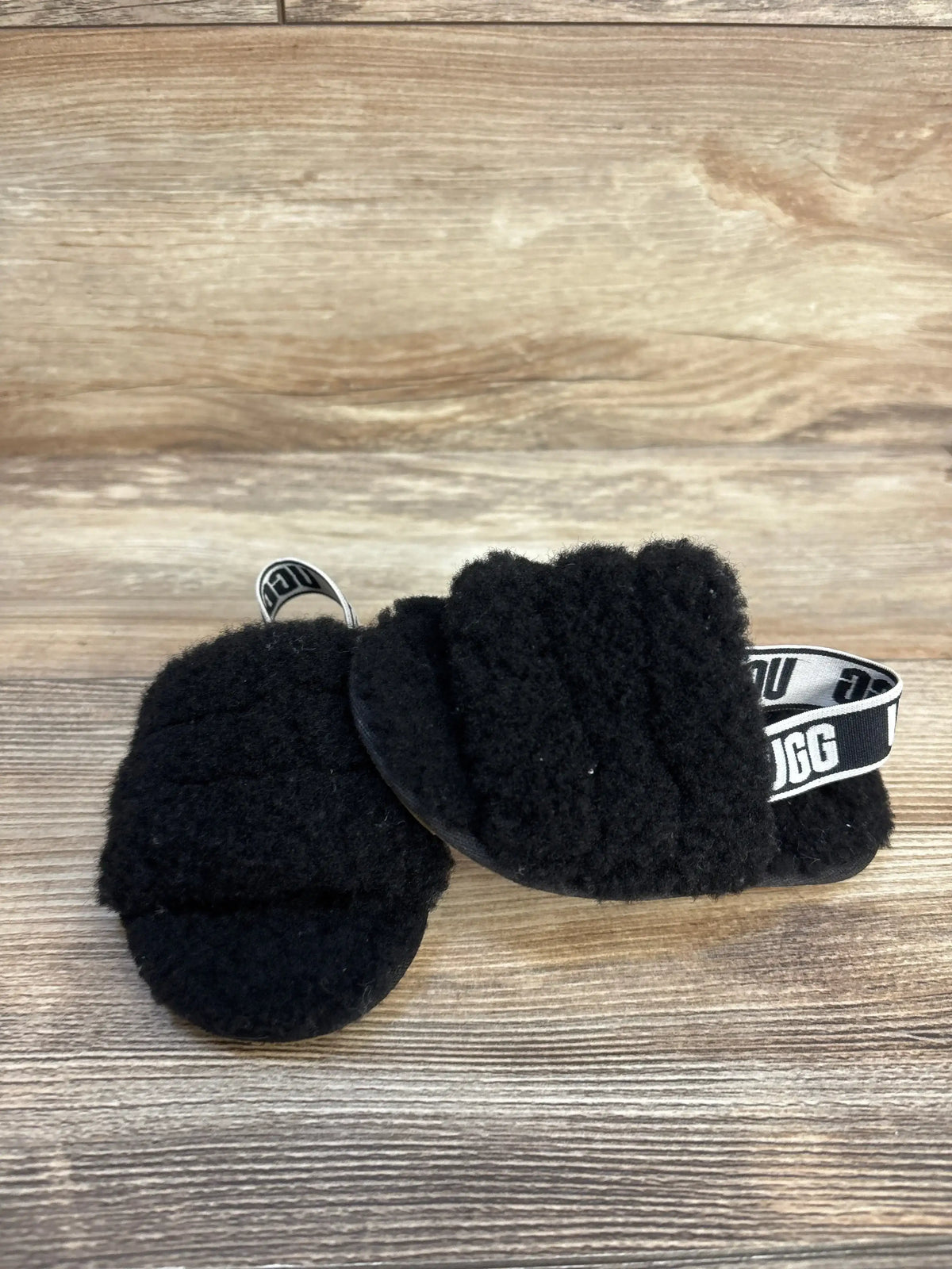 Angled front view of UGG Fluff Yeah slides showcasing black fluffy upper and branded elastic heel strap on wood background