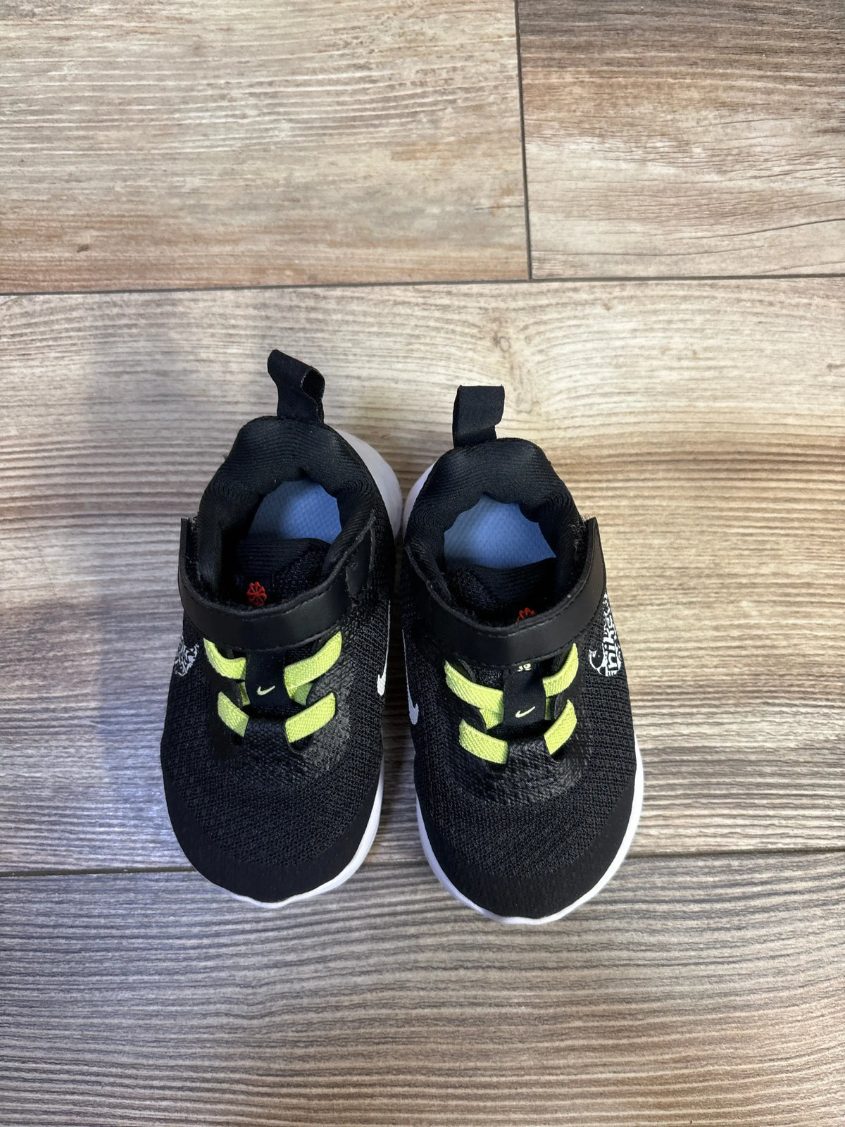 Top-down view of toddler Nike Revolution 6 sneakers with black knit upper, lime green elastic straps, white sole on wood floor, size 4C