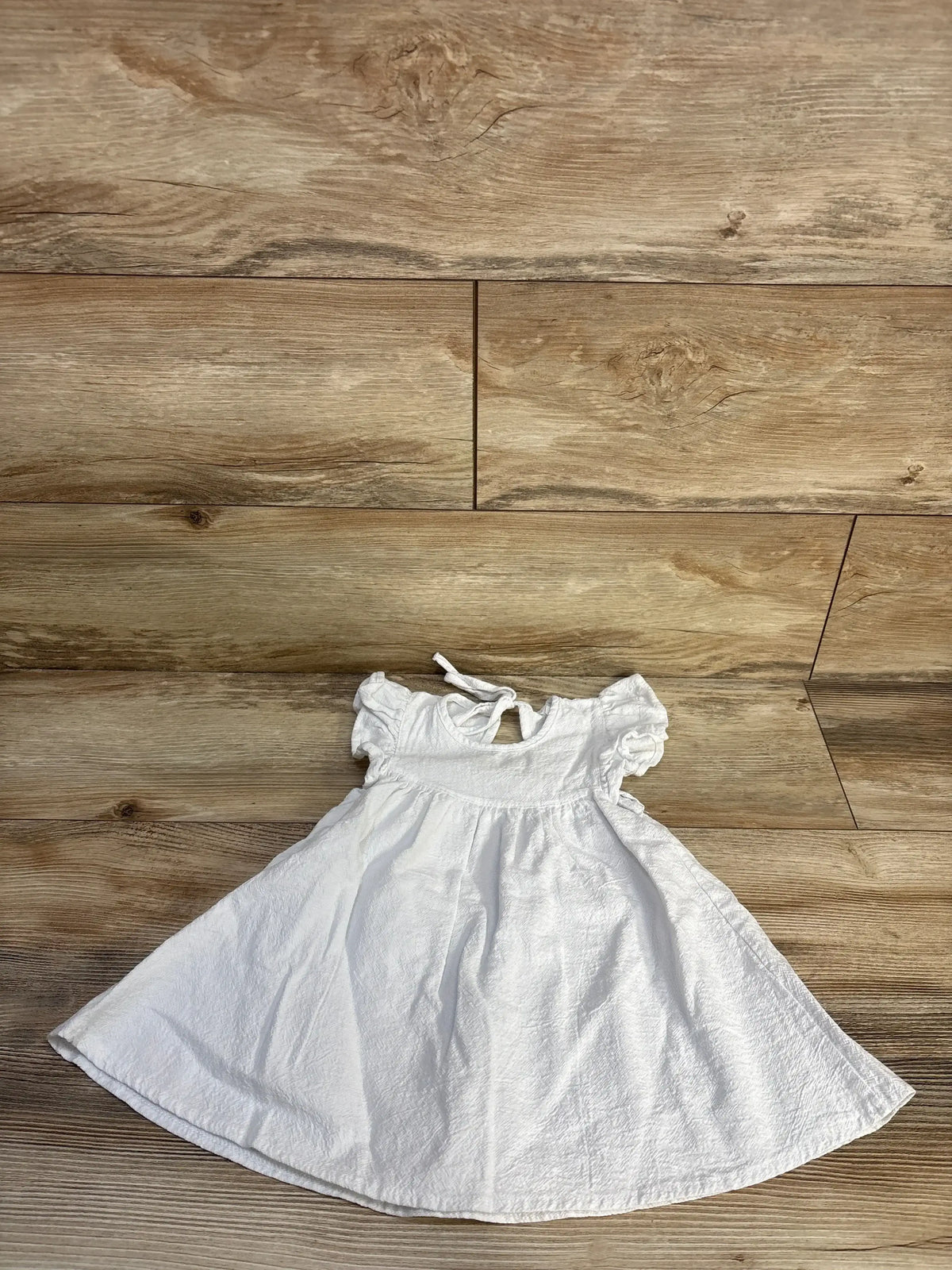Flat-lay of white textured flutter-sleeve baby dress 18 months showing tie-back keyhole and gathered skirt on wood plank floor