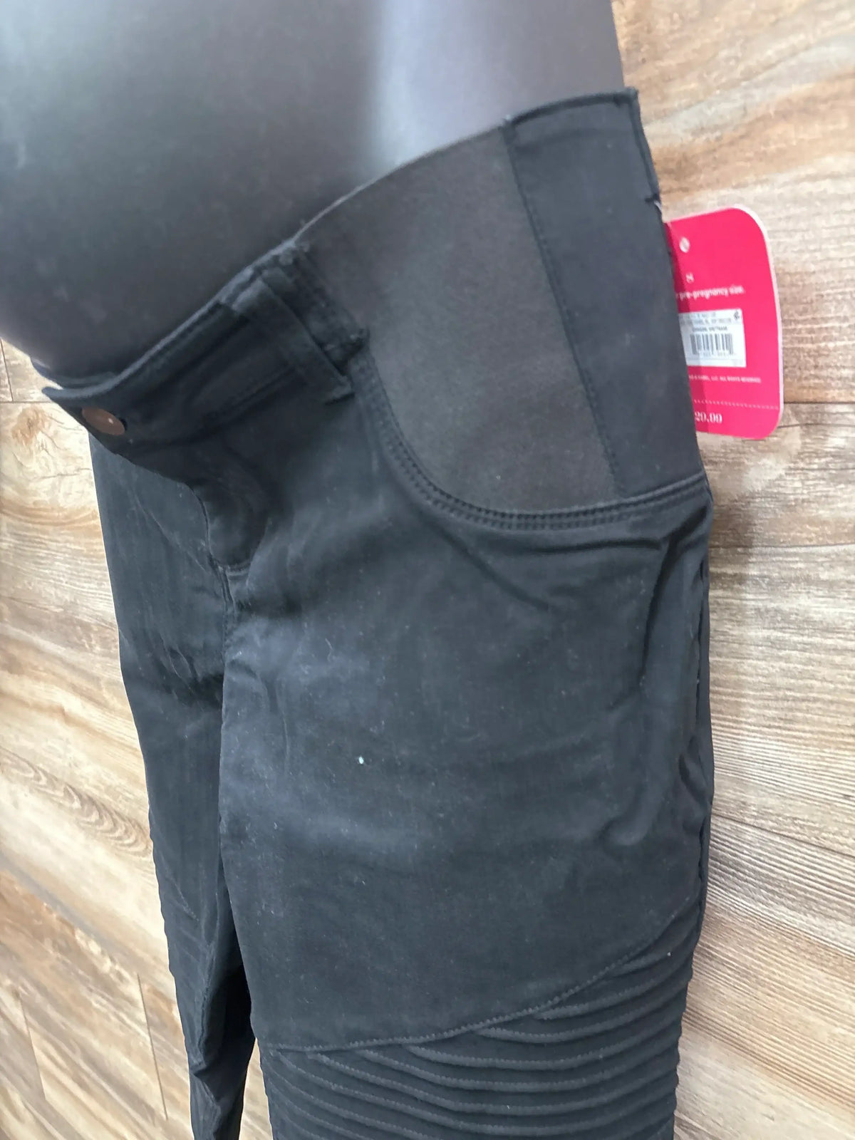Close-up side view of black maternity utility jeggings showing elastic over-belly panel, front pocket and ribbed knee detail on wood floor background