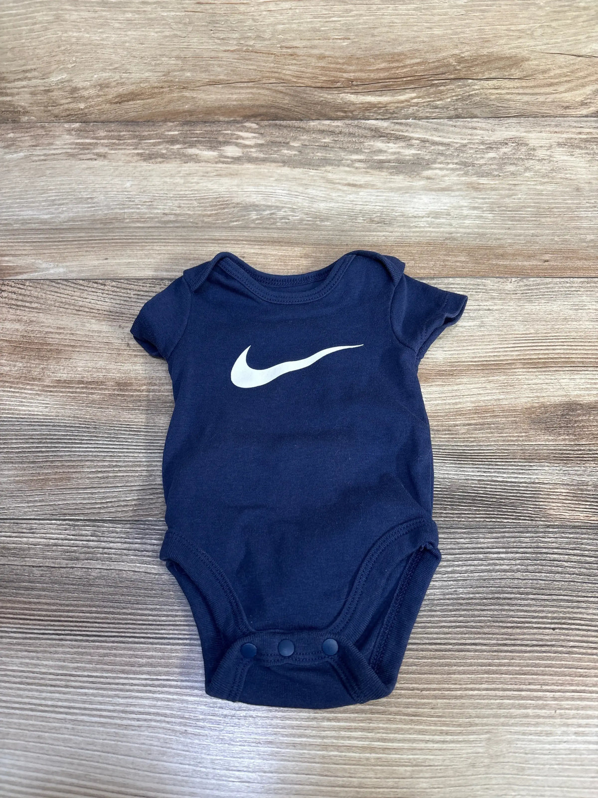 Flat lay of navy blue Nike baby bodysuit showcasing white swoosh logo and three snap closures on a wooden surface