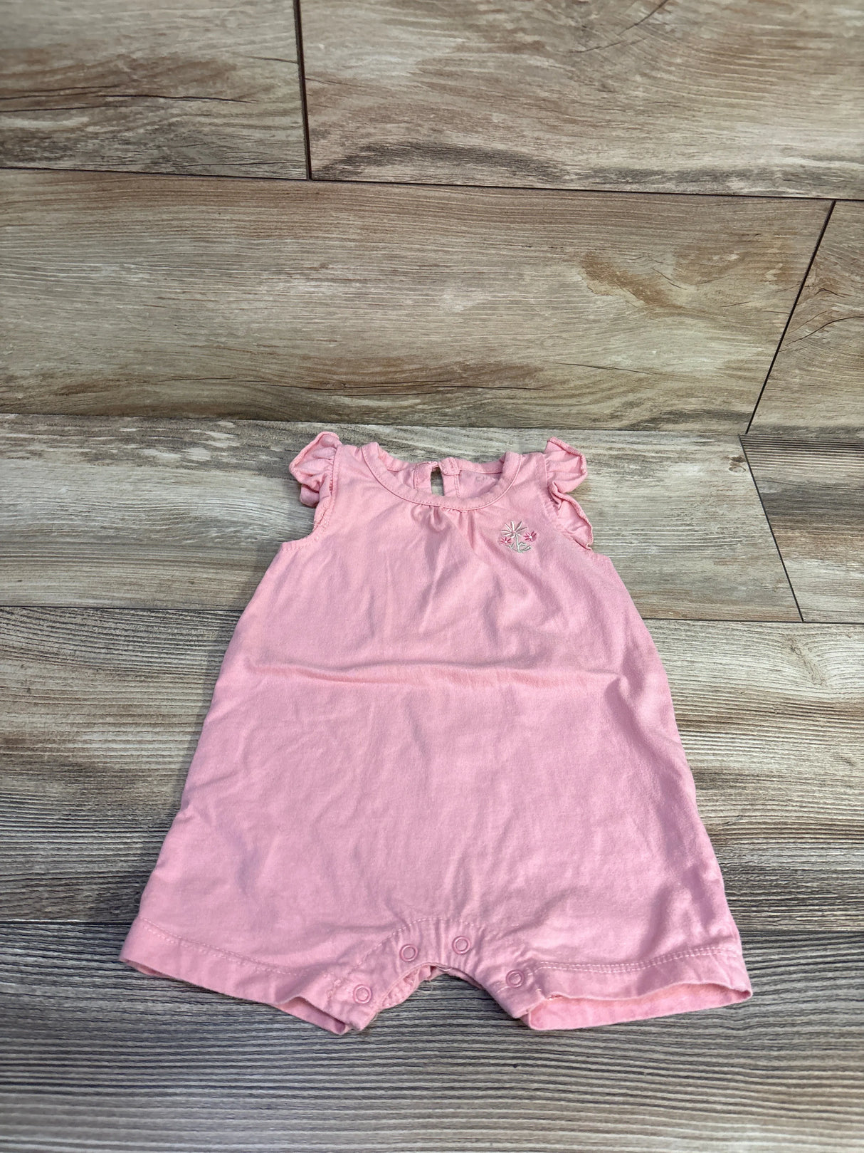 Flat lay of pink shortie romper, 6 to 9 months, showing ruffle sleeves, embroidered chest flower and leg snaps on wood floor