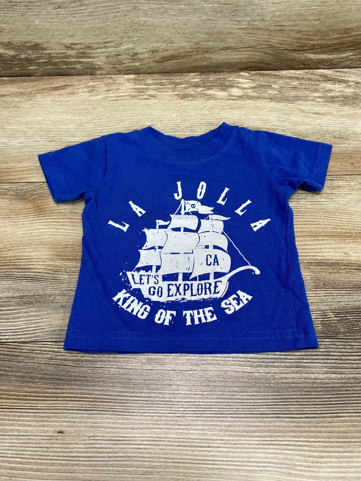 Flat lay front of blue Cotton Heritage 6 month baby shirt showcasing white sailboat graphic and the words La Jolla, Let’s Go Explore, and King of the Sea on a wood background