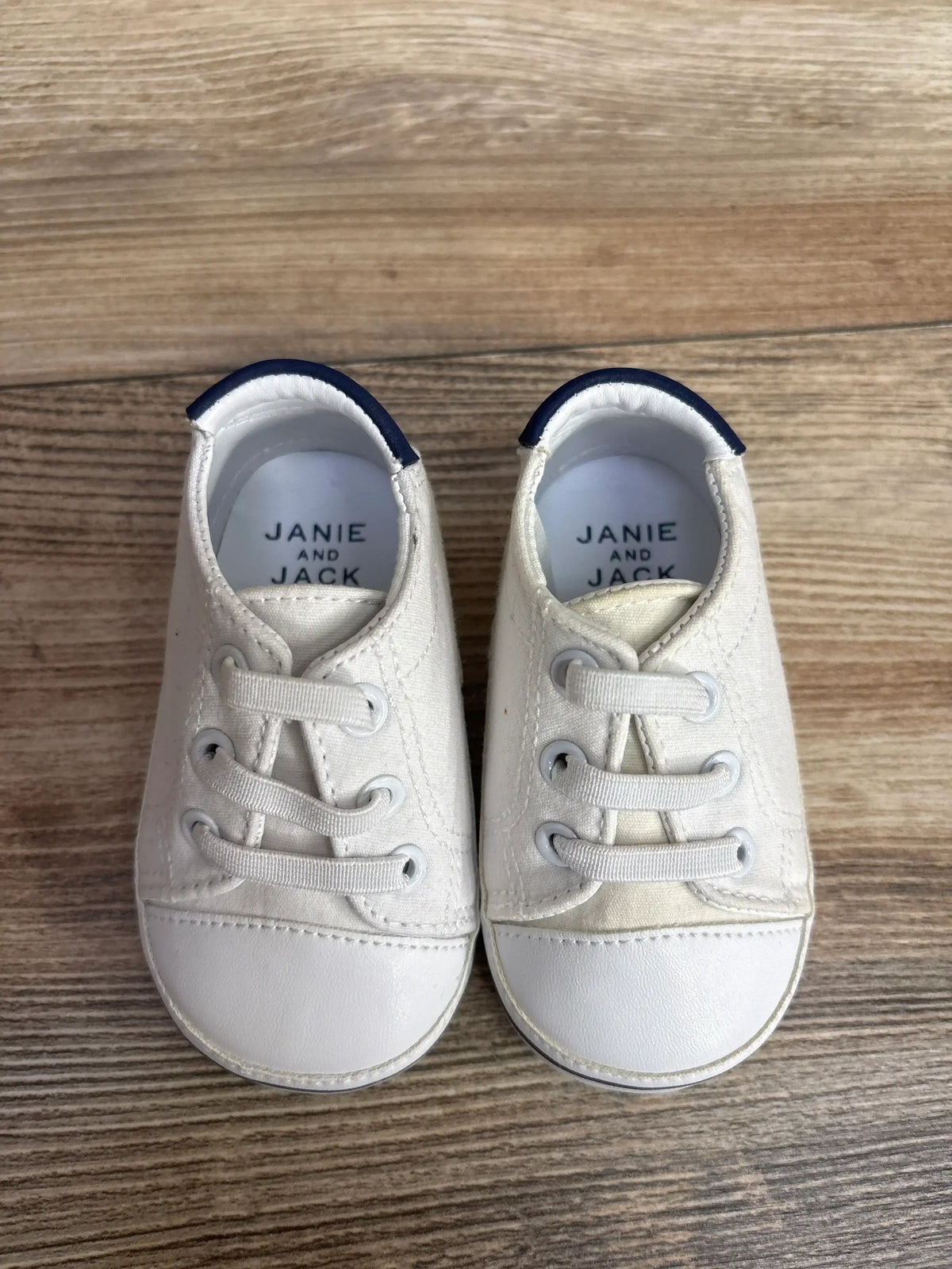 White Janie and Jack baby shoes with blue trim on wooden floor