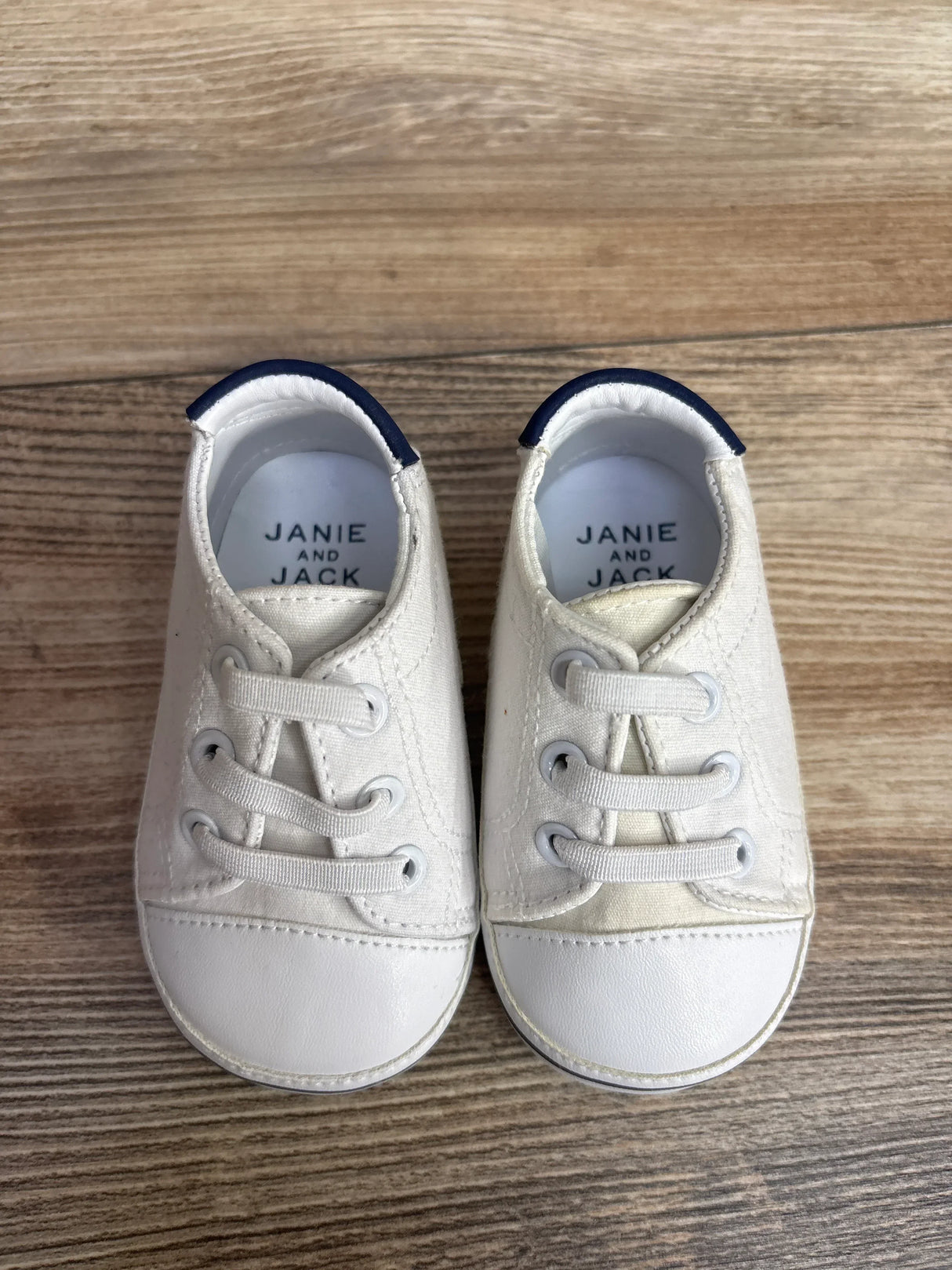 White Janie and Jack baby shoes with blue trim on wooden floor