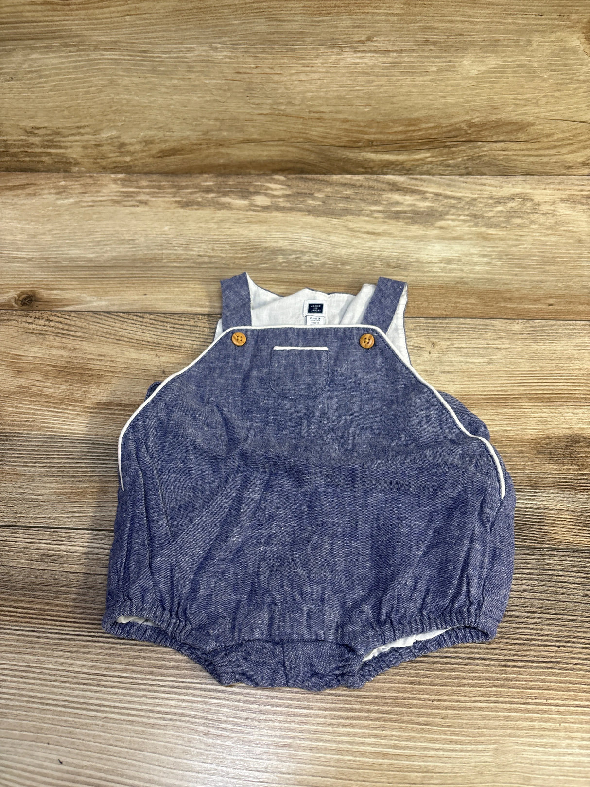 Top down view of chambray baby romper showing wooden button straps, white piping, and elastic leg openings on wood background