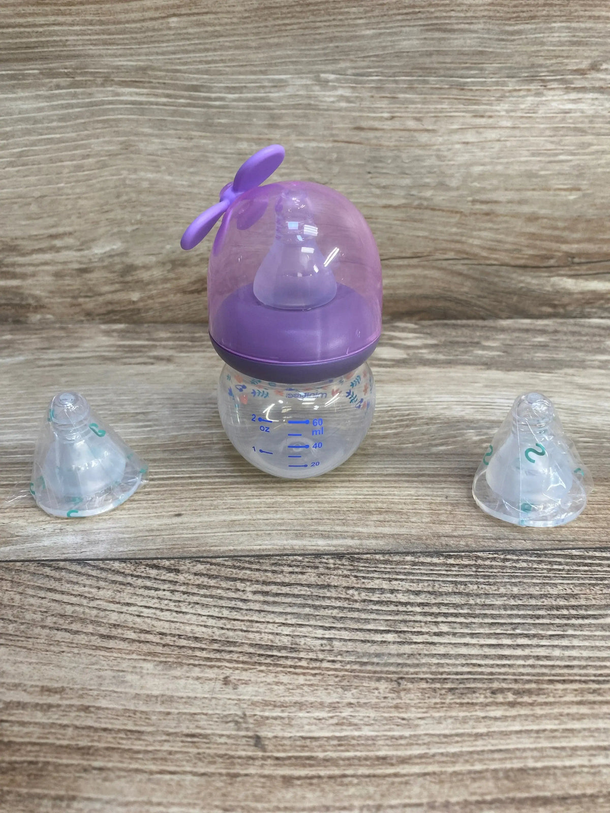 Front view of MINITree 2oz purple baby bottle with dome cap and visible 2 oz/60 ml markings, plus two sealed extra nipples on wood background