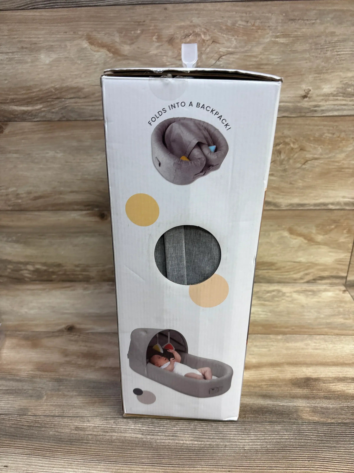 Portable baby lounger with canopy, folds into a backpack, shown in packaging on wood floor