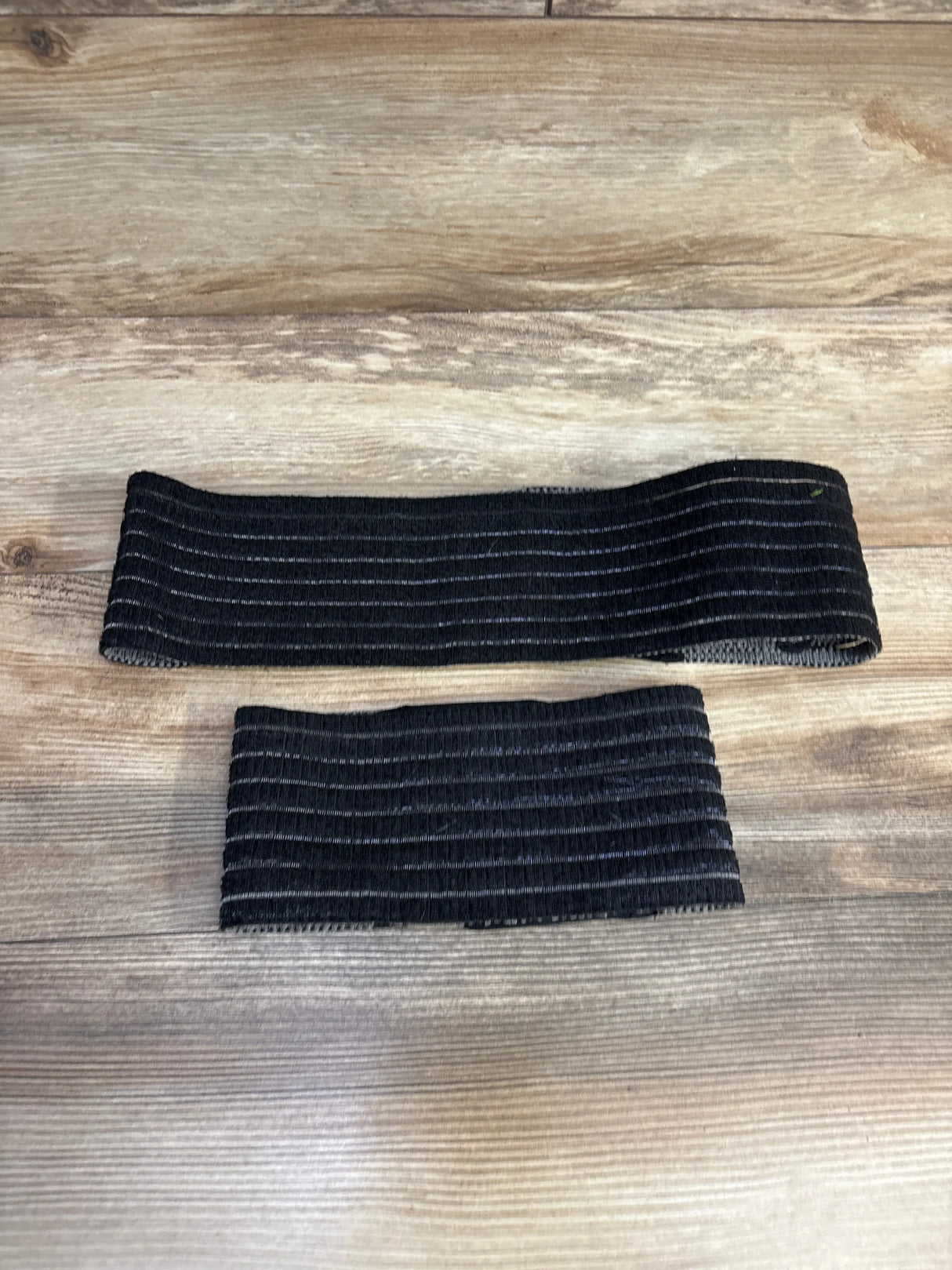 Top-down view of Belly Bandit 2-in-1 Hip Bandit (black, XS–M) showcasing a wide hip support band and a narrow compression strap on a wood floor