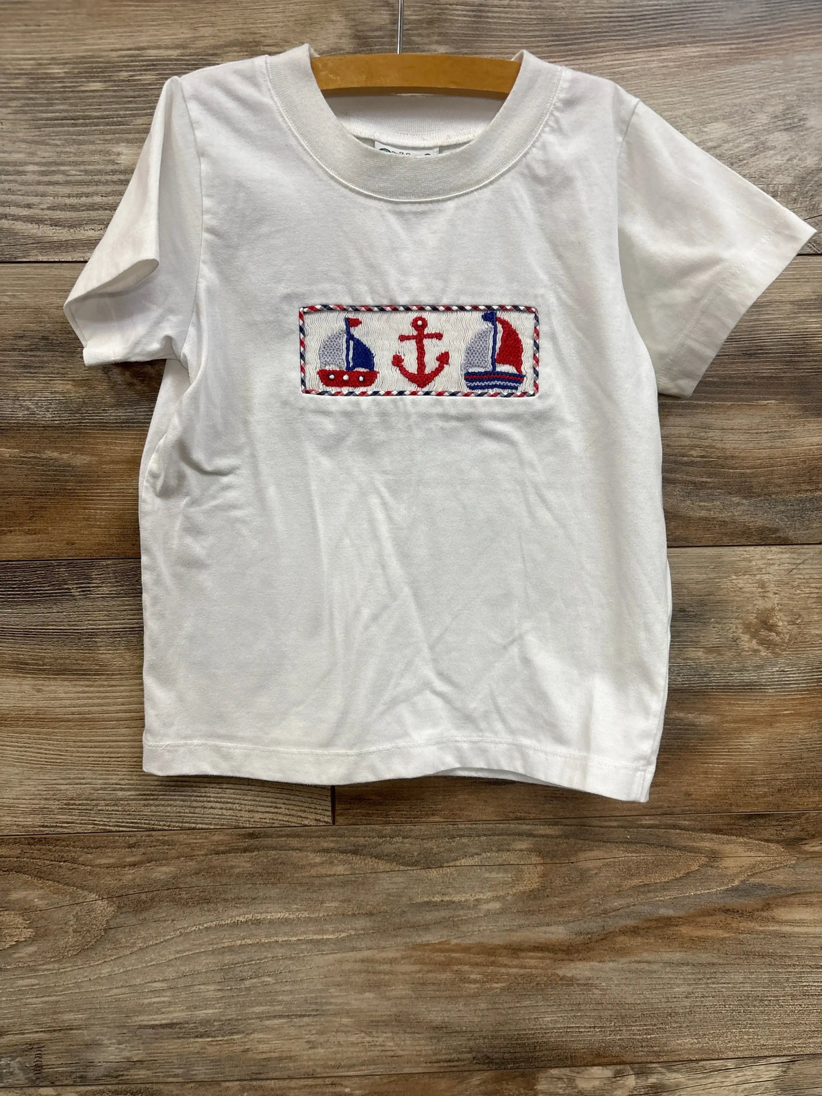 Front view of white toddler smock shirt with smocked panel featuring two sailboats, red anchor, and red-blue braided trim on wooden background