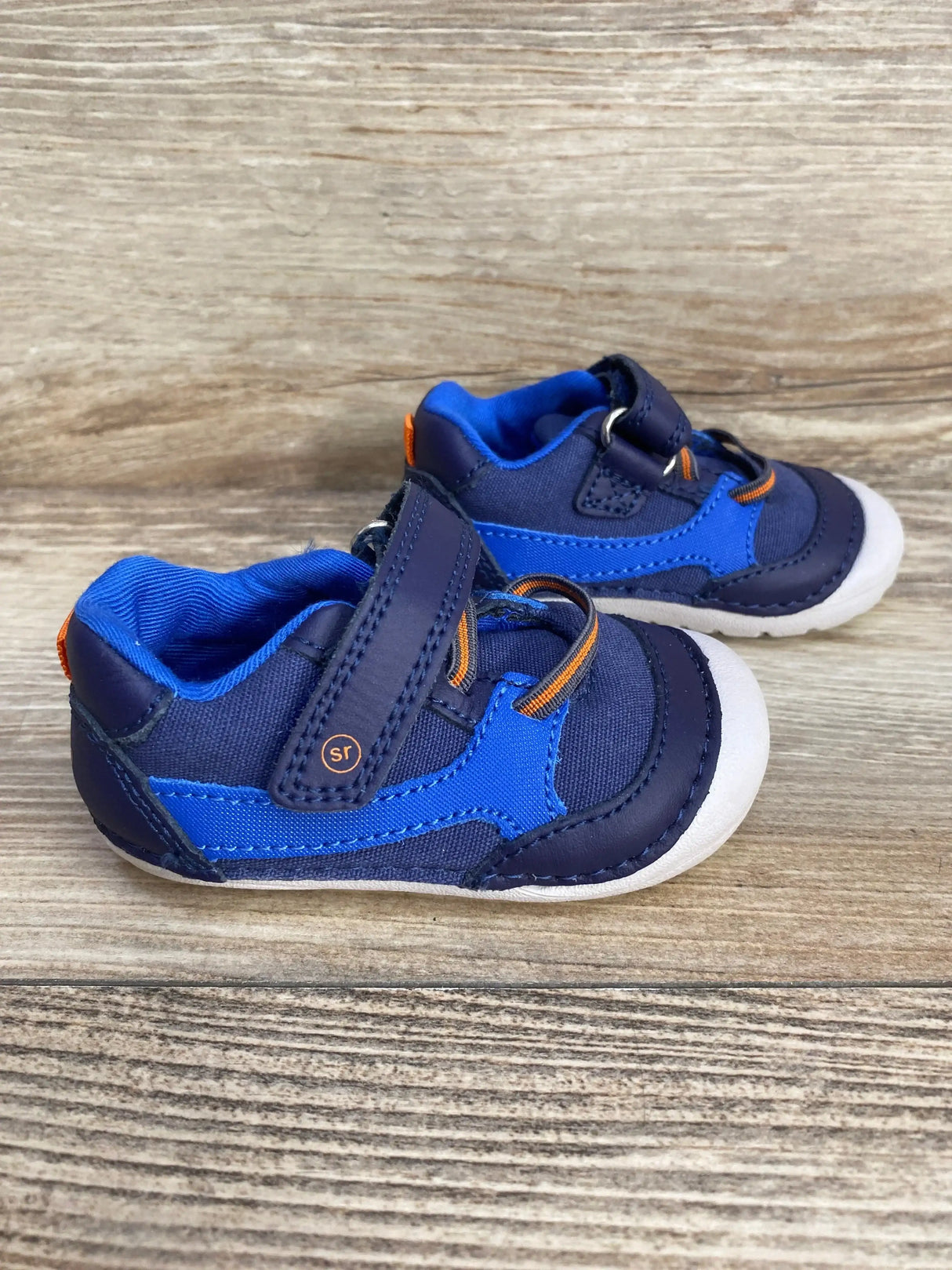 Side view of Stride Rite Soft Motion Kylin toddler sneakers in blue showing velcro strap, cushioned collar, and white rubber toe on wood background