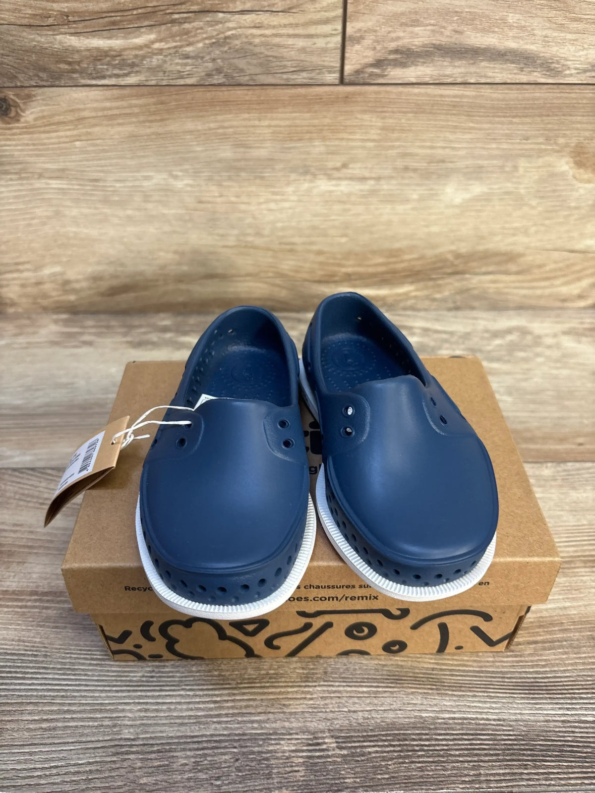 Navy blue slip-on kids shoes with white soles displayed on a shoe box, wood background