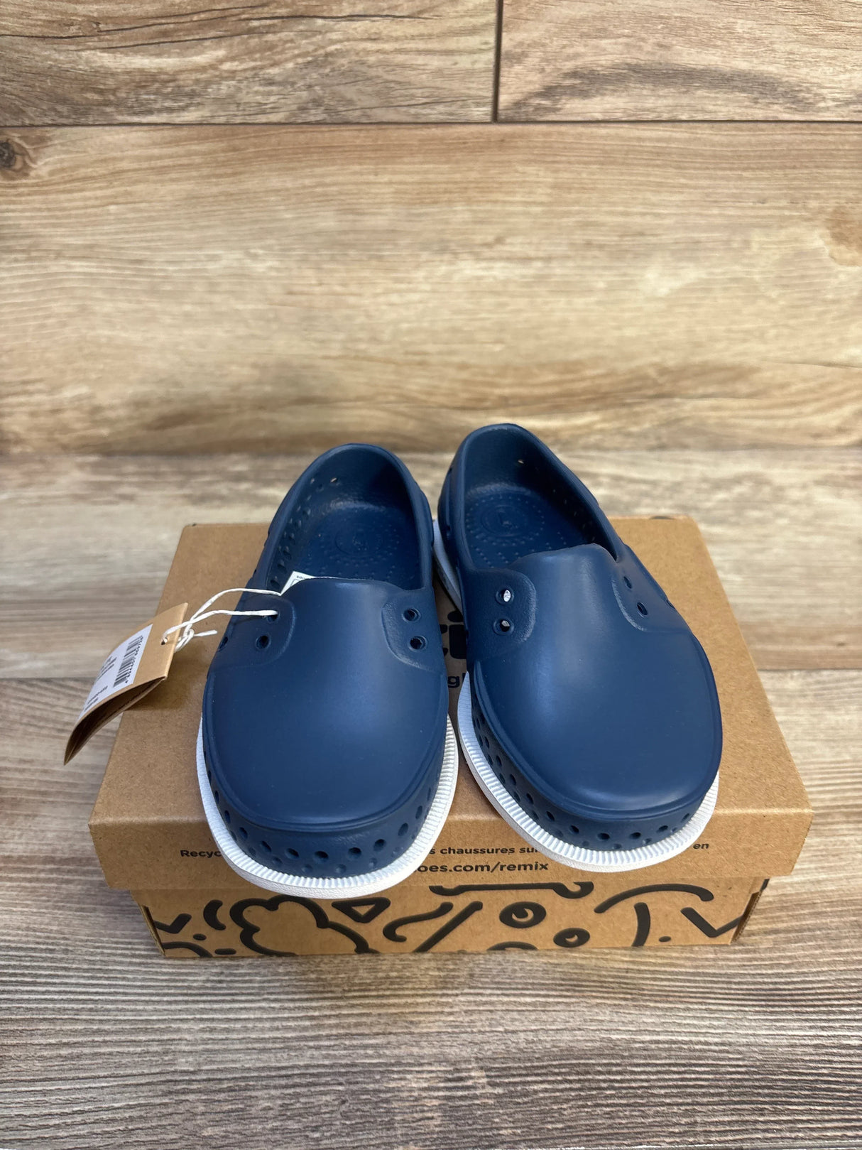Navy blue slip-on kids shoes with white soles displayed on a shoe box, wood background
