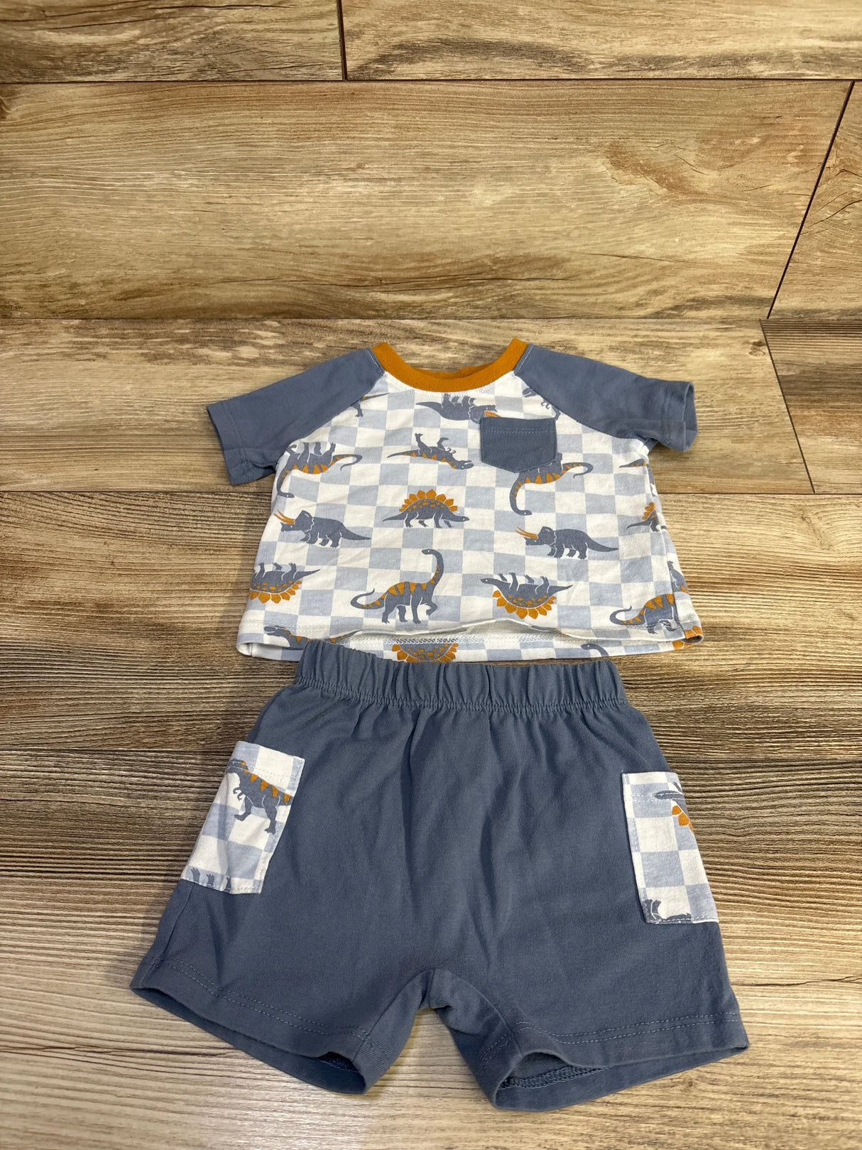 Flat lay of blue 2-piece dinosaur shirt and shorts showing checkerboard dino print, chest pocket and elastic waist on wood