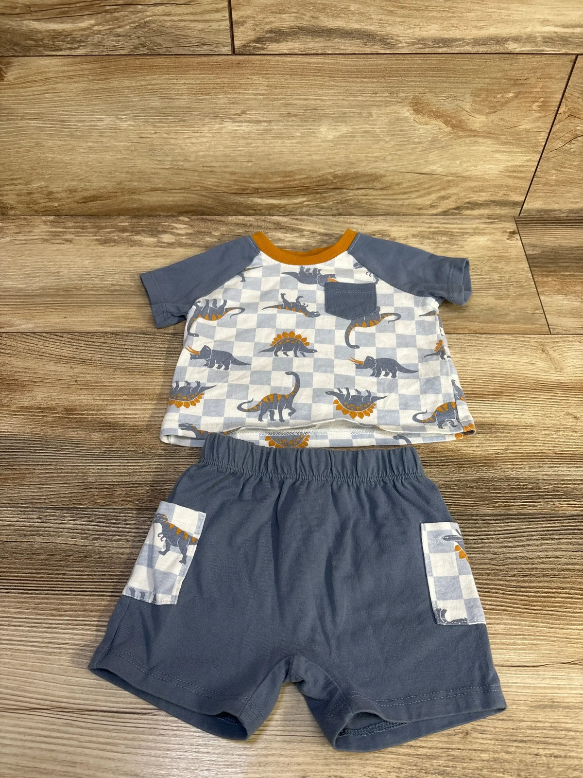 Flat lay of blue 2-piece dinosaur shirt and shorts showing checkerboard dino print, chest pocket and elastic waist on wood