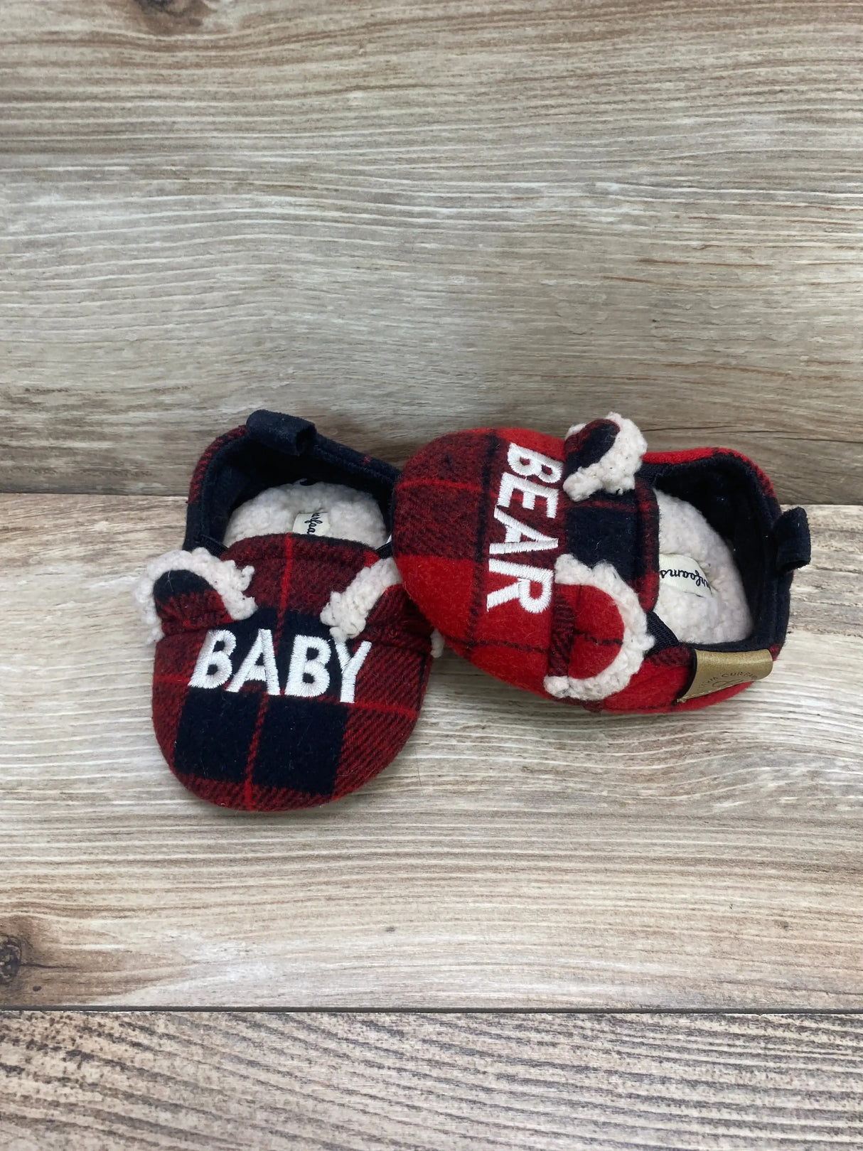 Top-down of Dearfoams baby slippers red and black plaid with BABY and BEAR embroidery and faux shearling lining on wood