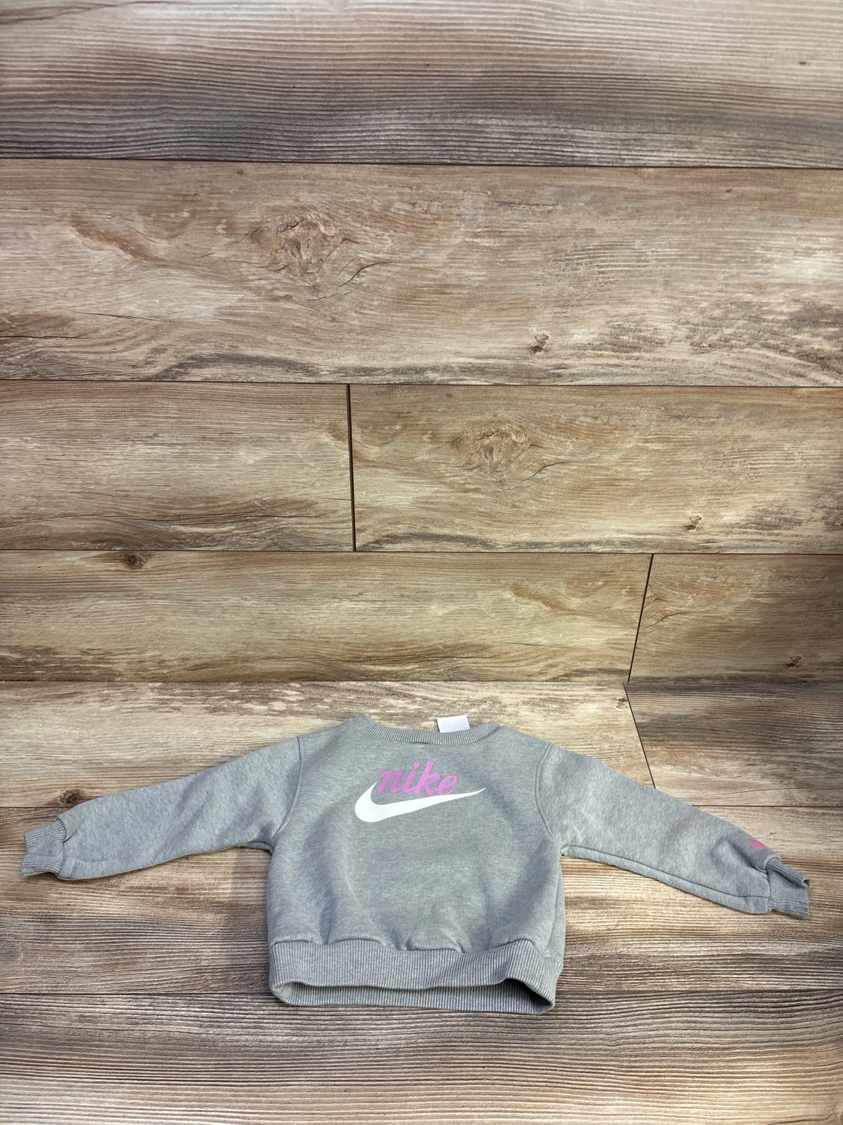 Flat lay of grey toddler Nike pullover sweatshirt size 18 months showcasing pink Nike script and white swoosh on a wood plank background