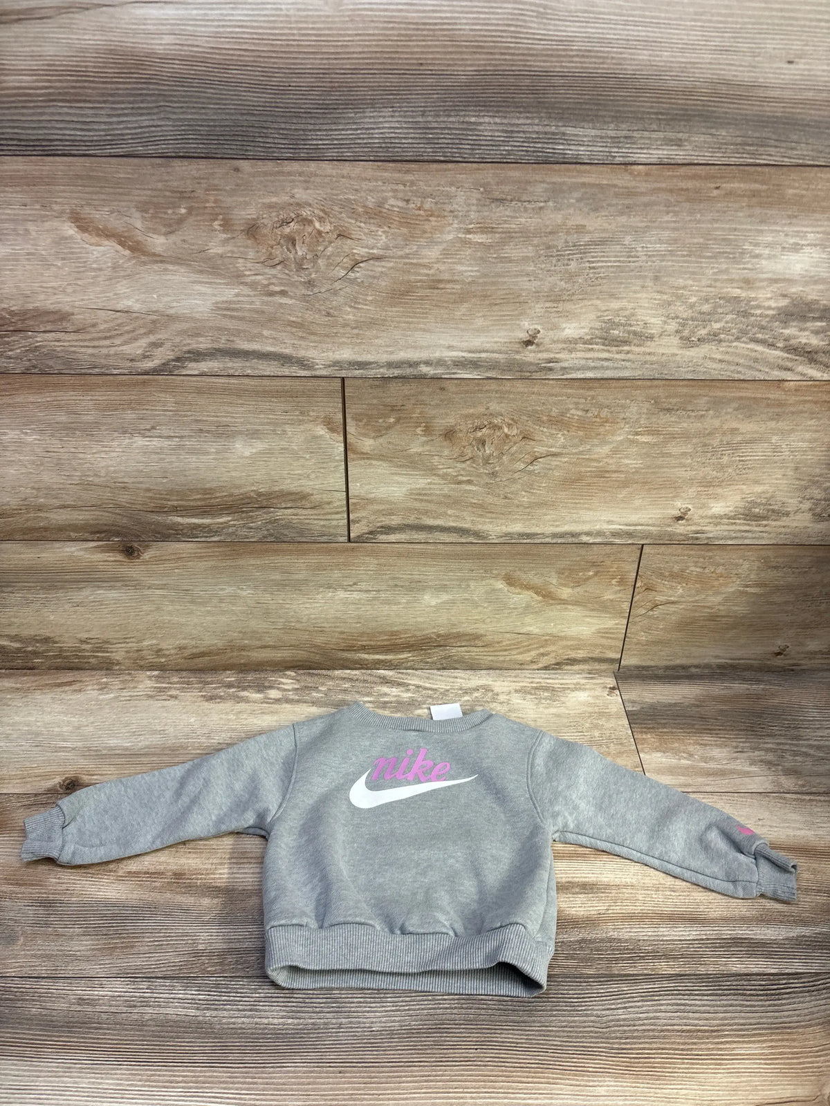 Flat lay of grey toddler Nike pullover sweatshirt size 18 months showcasing pink Nike script and white swoosh on a wood plank background