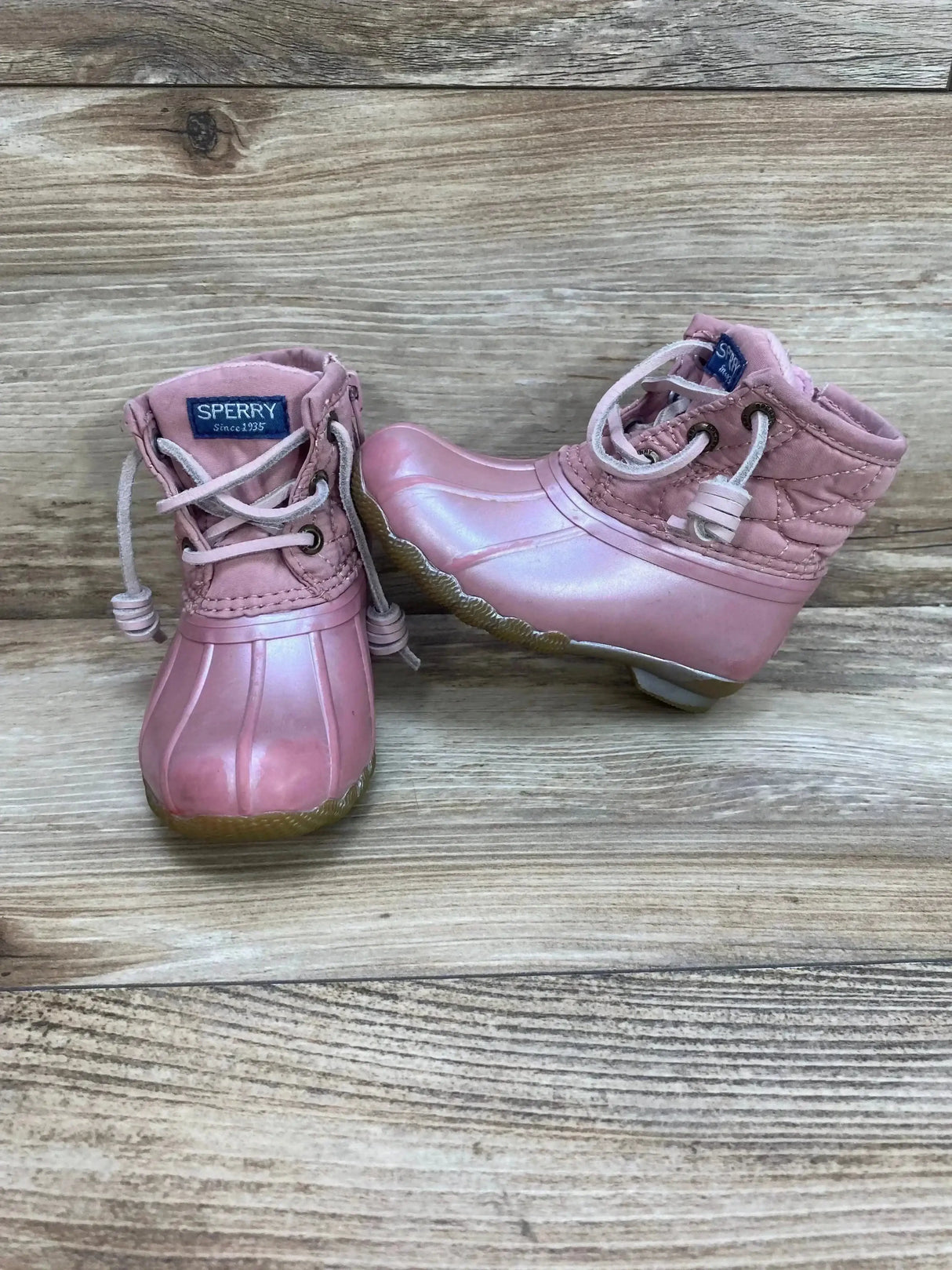 Three-quarter view of toddler Sperry Saltwater pink duck boots showing quilted ankle, lace ties, rubber toe and gum sole on wooden plank background