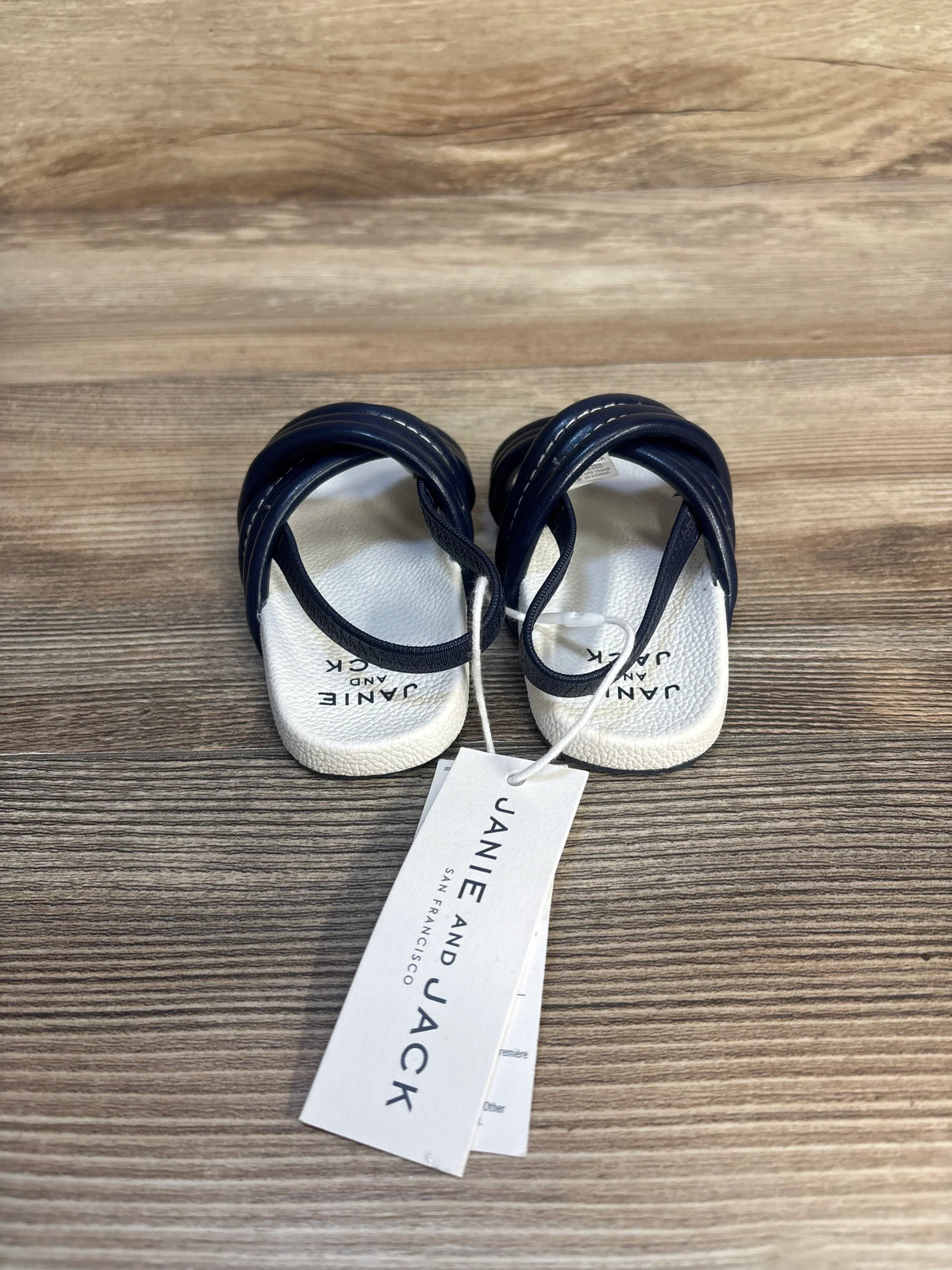 Overhead of Janie and Jack navy cross-strap baby pool slides showing crisscross straps and brand tag on wood surface