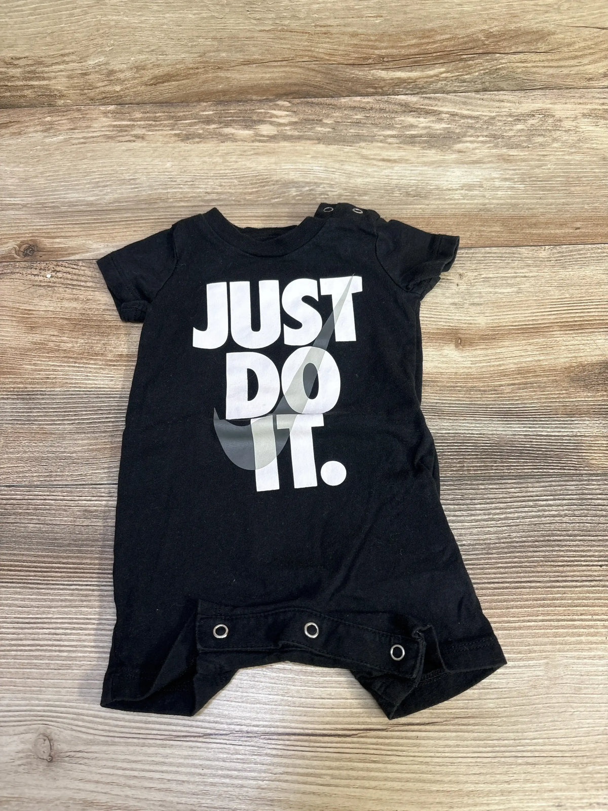 Flat lay of black Nike shortie romper showing large white JUST DO IT print and three snap leg closures on a wood floor