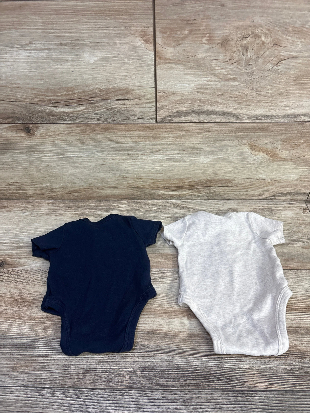 Flat lay of two newborn Henley bodysuits (navy and light gray) showcasing short sleeves and snap crotch with wood plank background