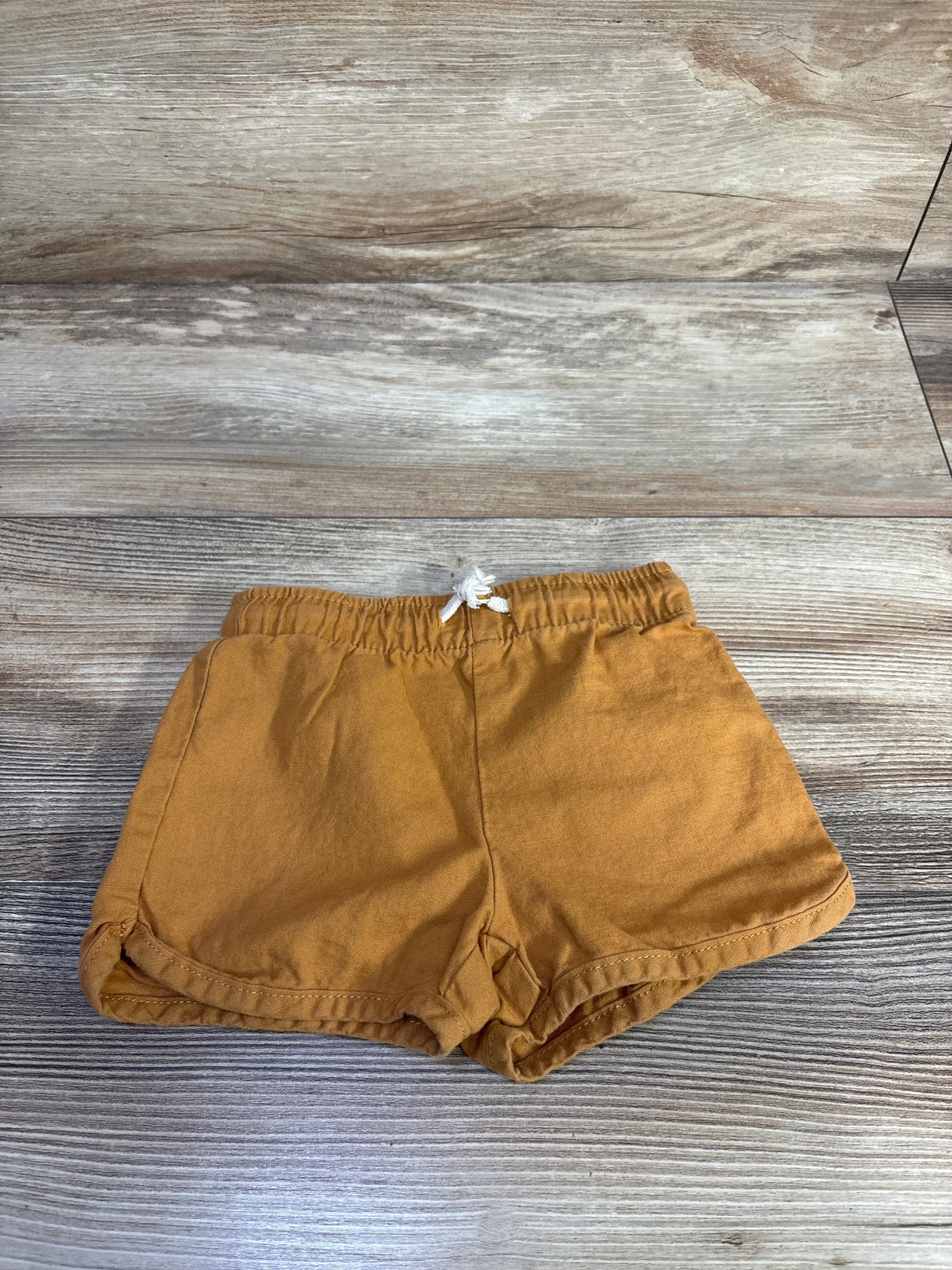 Flat-lay front of Little Co. caramel organic baby shorts showing elastic waistband with white drawstring on wood background