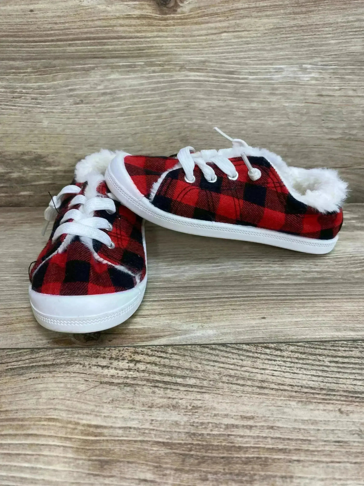 Front three quarter view of toddler slip-on sneakers showcasing red and black buffalo plaid exterior, white faux fur lining and white rubber sole on wood background