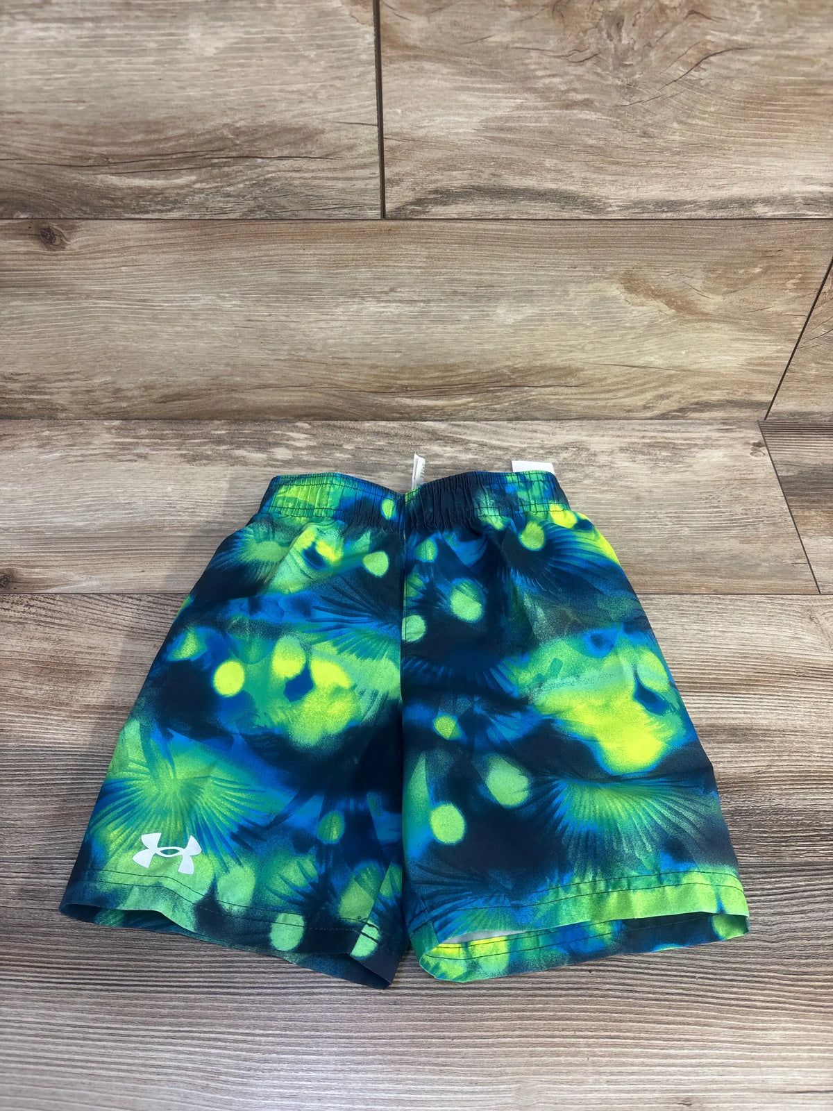 Front view of Under Armour navy swim trunks in size 4T with a green and blue tropical pattern on a wood floor
