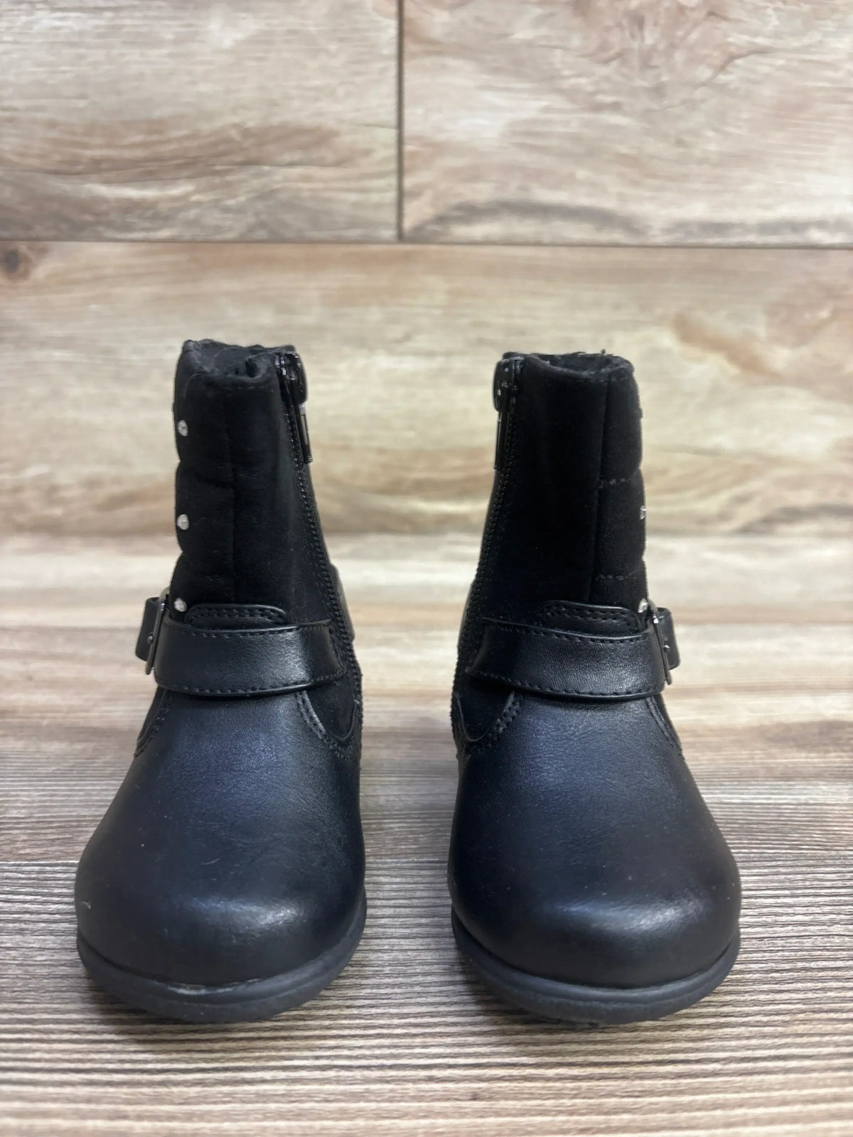 Pair of black toddler boots with side zippers and decorative straps on wood surface