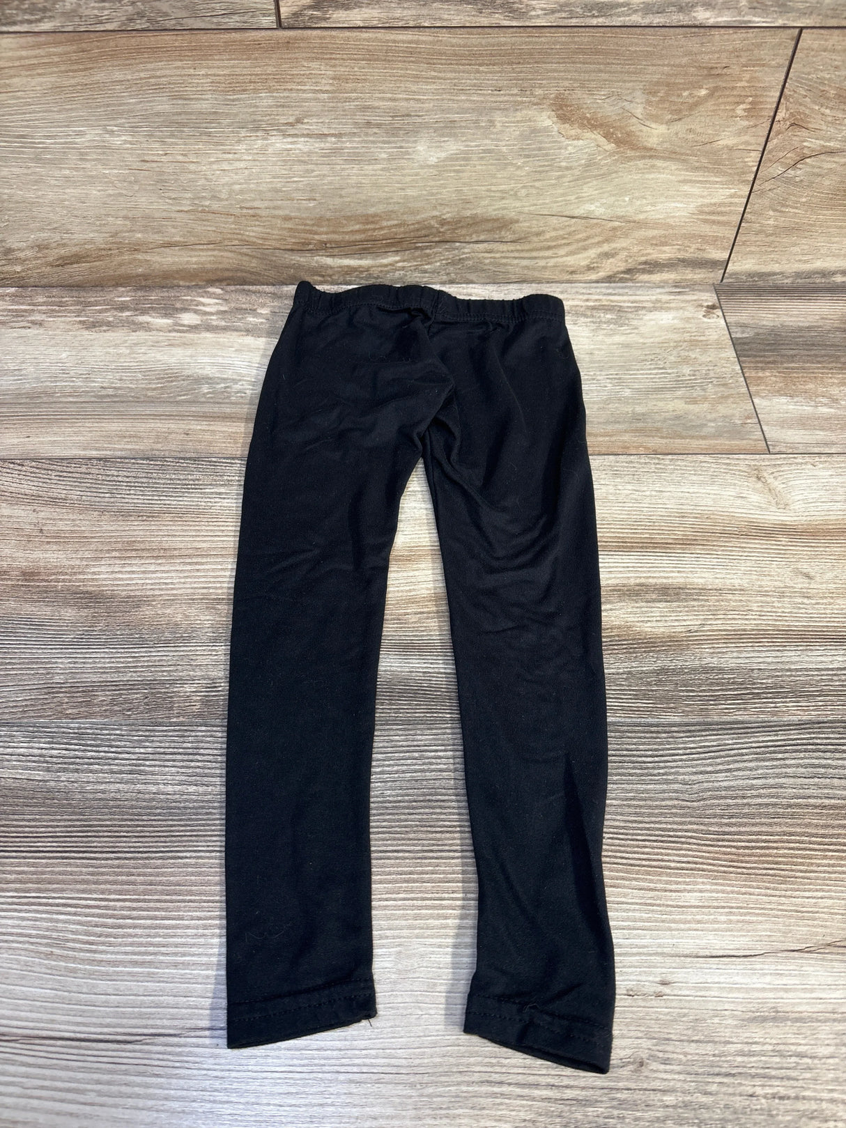 Flat lay of black toddler leggings size 2T showing elastic waistband and straight slim legs on light wood floor background