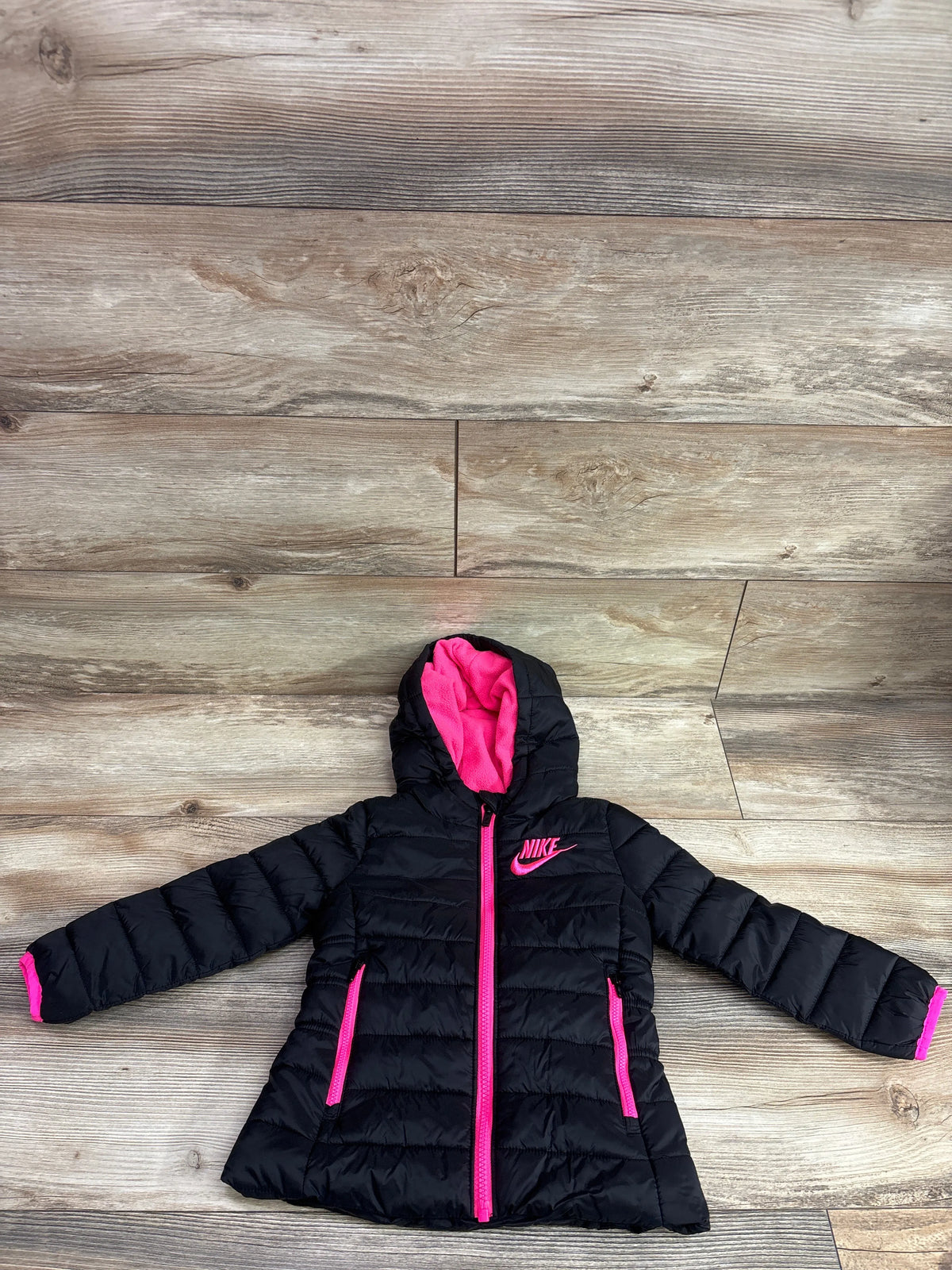 Top view of black Nike hooded puffer jacket, size 2T, showcasing pink lining, zipper, trim, and quilted panels on wood floor