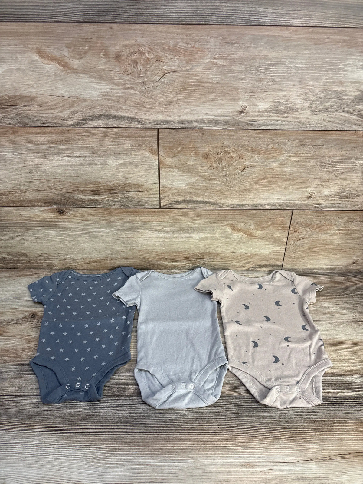 Top view of Cloud Island 3-pack grey bodysuit set showcasing star, solid, and moon prints on a wood-look background