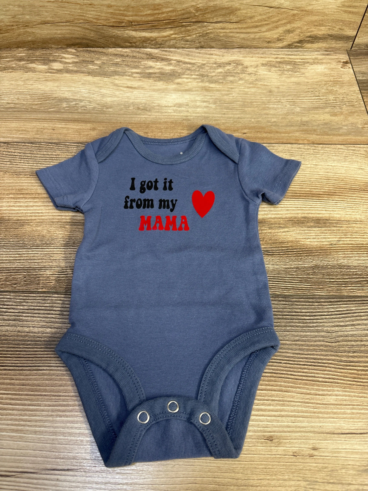 Flat lay of blue Hanes infant bodysuit (0-6M) showcasing I got it from my MAMA text and red heart on wood background