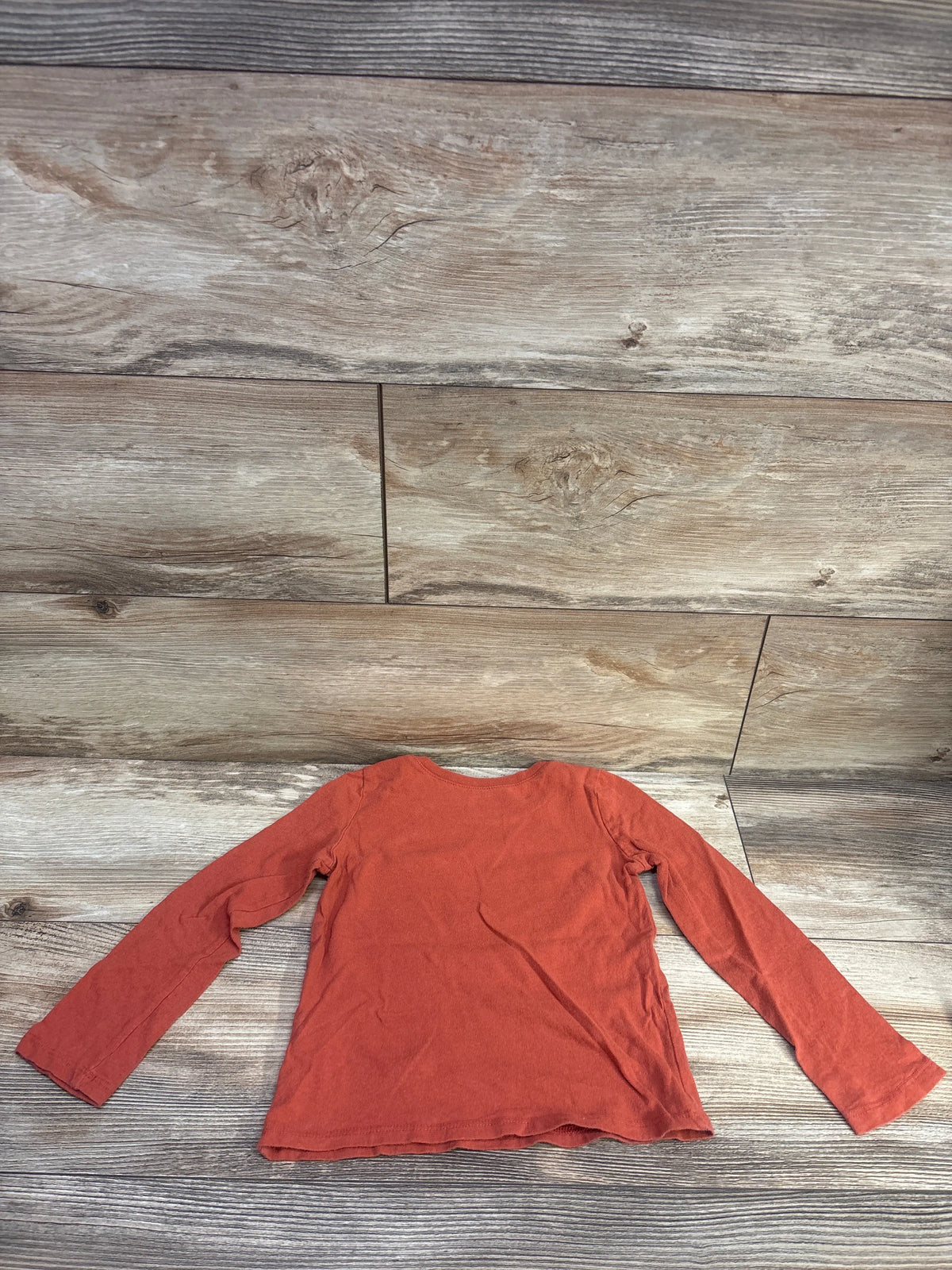Top view of a rust long-sleeve children’s shirt laid flat on a wood-grain background